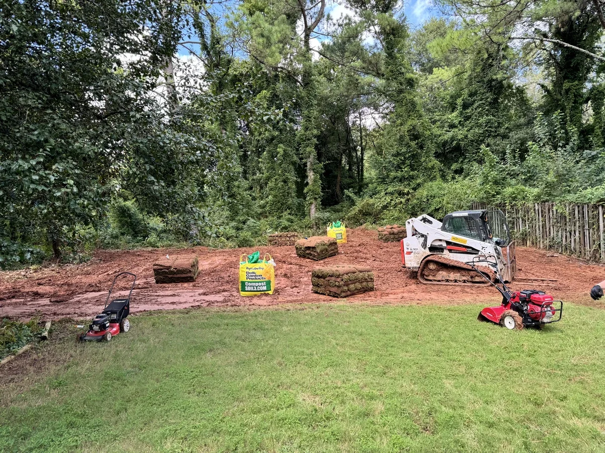A backyard scene with a partially dug area, stacks of sod, and yard equipment including a mini bulldozer, a grass roller, and bags of Garden Products soil. There are trees and a wooden fence in the background.