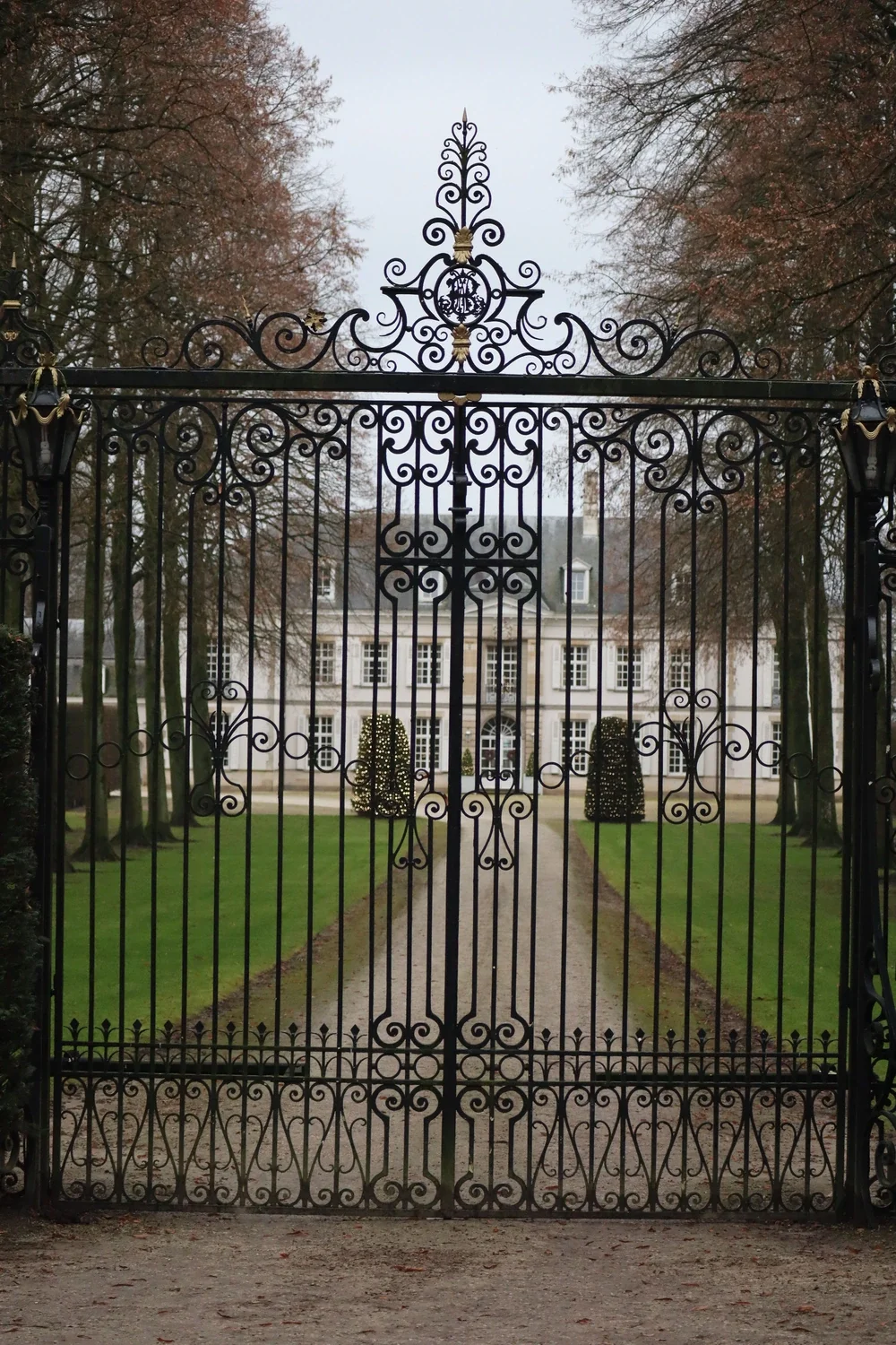 An ornate black wrought iron gate with intricate scrollwork, leading to a large, elegant mansion with multiple windows and a manicured lawn, surrounded by tall trees on an overcast day.