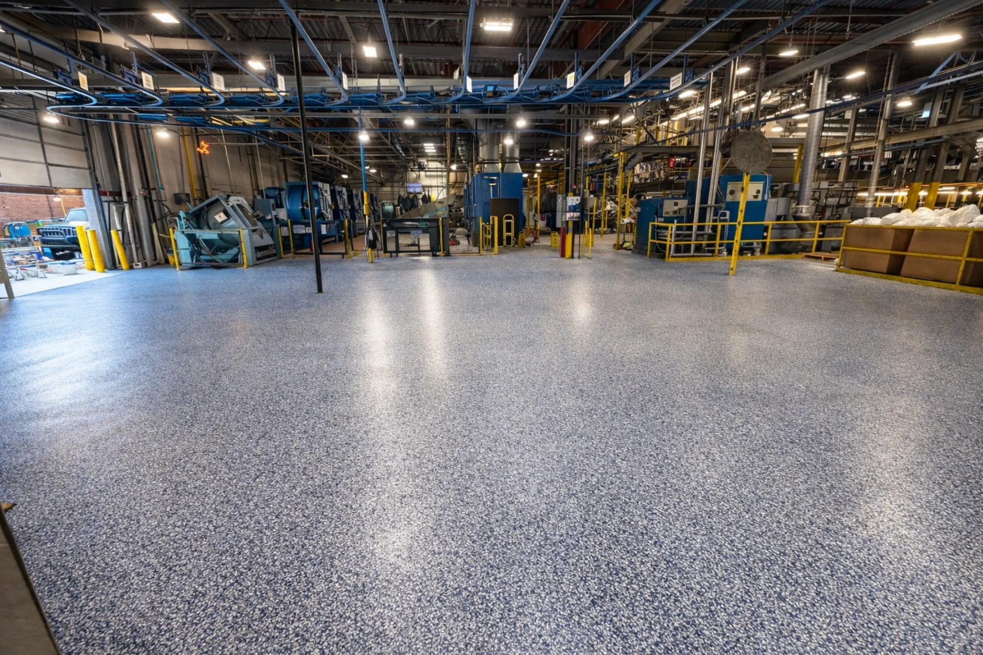 An new floor in an industrial space with yellow safety barriers, mechanical equipment, and exposed piping and wiring on the ceiling.