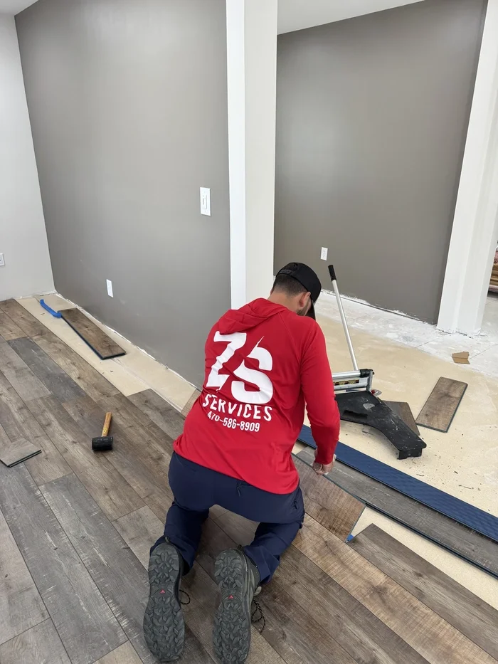 A worker in a red hoodie installing a wooden floor in a room under renovation. The worker is kneeling on the floor, using a flooring cutter to position wooden planks. The room has gray walls, some tools, and area of the floor still under construction.