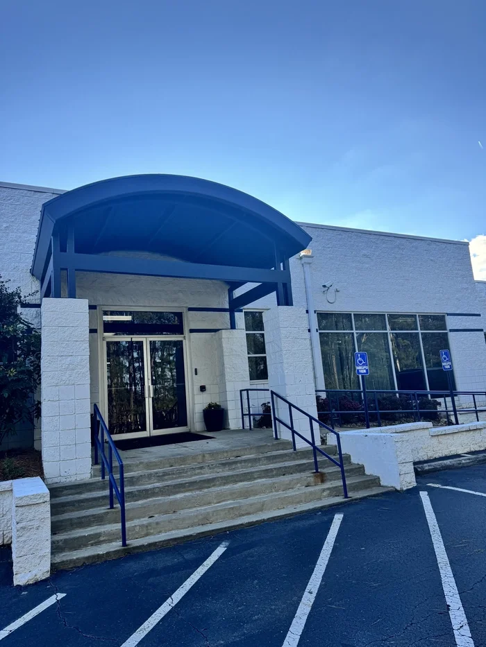 Exterior of a white building with a blue awning over the entrance, accessible ramp, and clear parking spaces with space for handicapped parking.