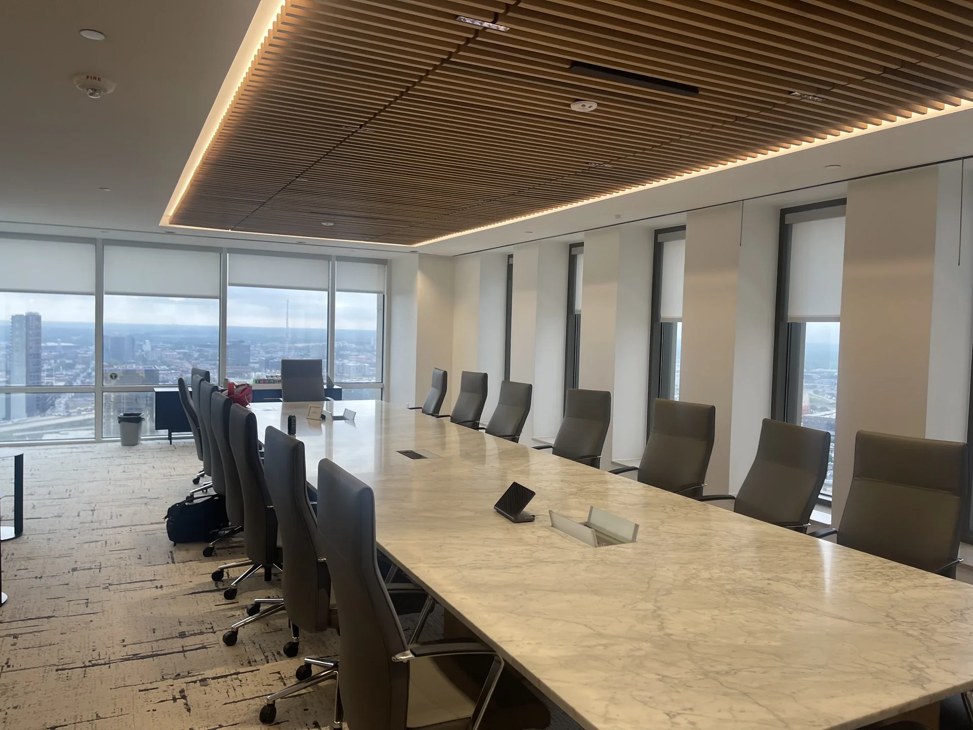 A modern conference room with a long table, black chairs, large windows showing city views, and a wooden slatted ceiling with built-in lighting.