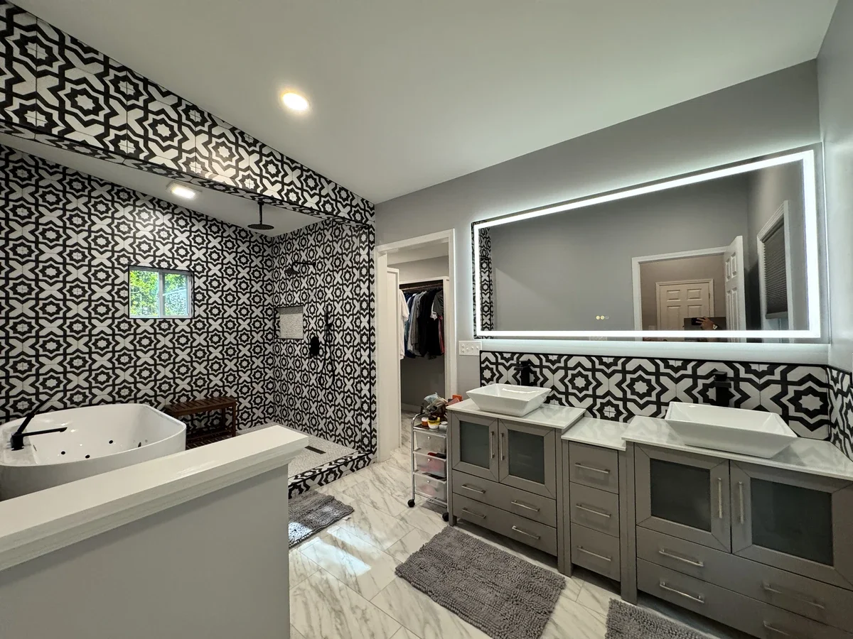 Modern bathroom with black and white patterned tile walls, a large mirror with LED lighting, double sinks, a vanity with drawers, a walk-in shower area with patterned tiles, a small window, and a bathtub.
