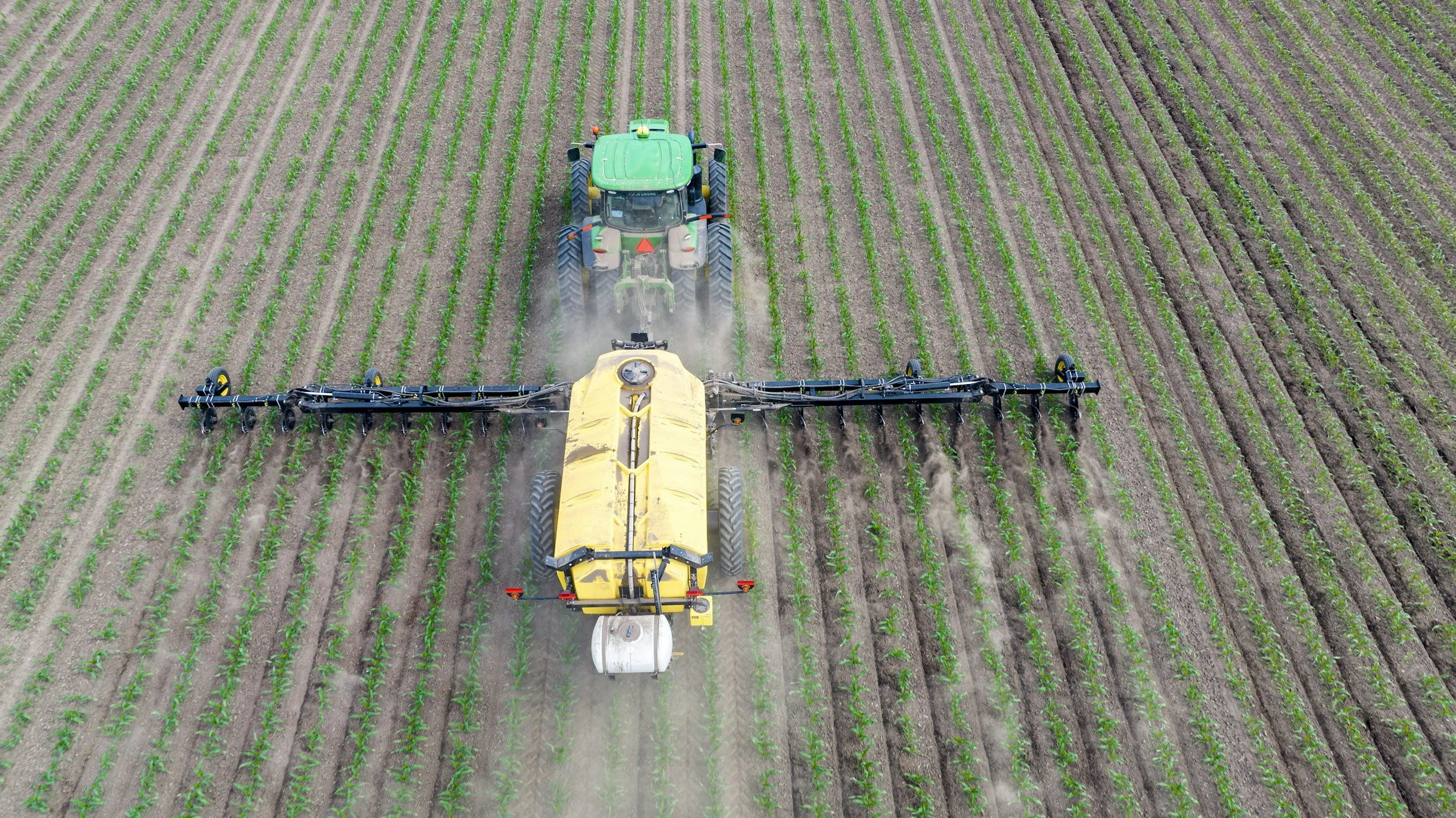 Aerial view of a tractor pulling a large spray tank, spraying crops in a vast green farm field in straight rows.