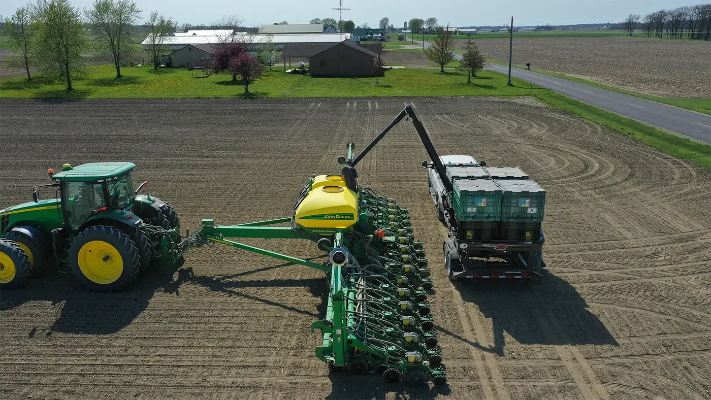 A green John Deere tractor is working on a farm field, pulling a large seed planter. It is transferring seeds into a smaller green seed container on a trailer, with a distant farmstead and open farmland in the background.