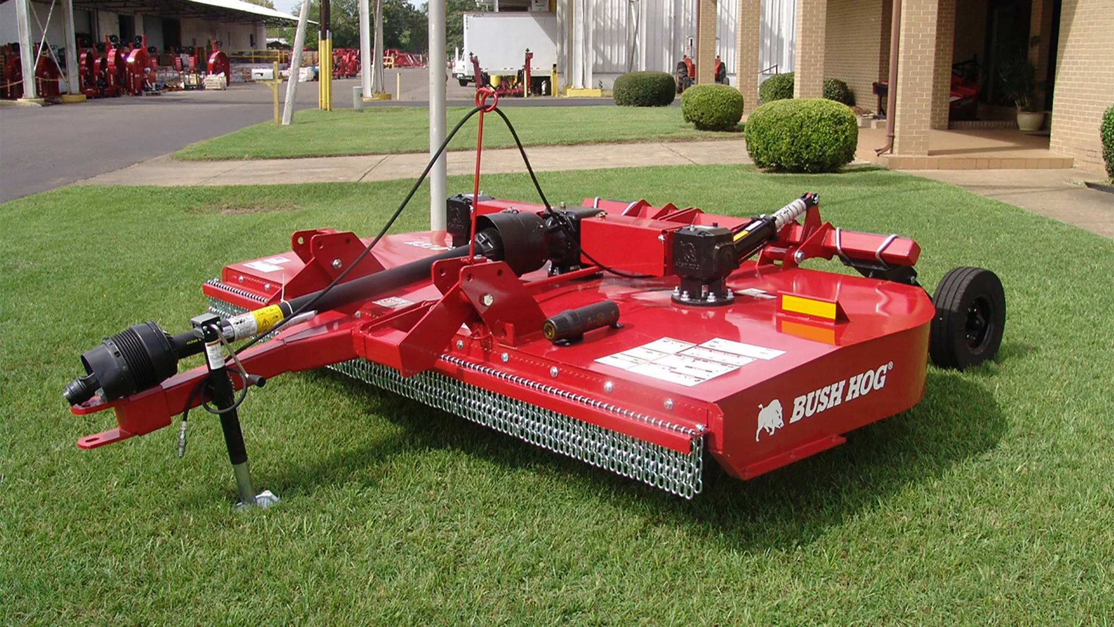 Red Bush Hog rotary cutter mower on a grassy area, with a building and sidewalk in the background.