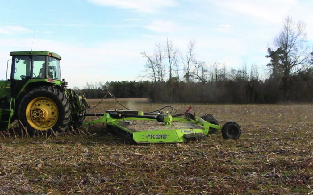 A green tractor pulling a large mower attachment in a field with leafless trees in the background.