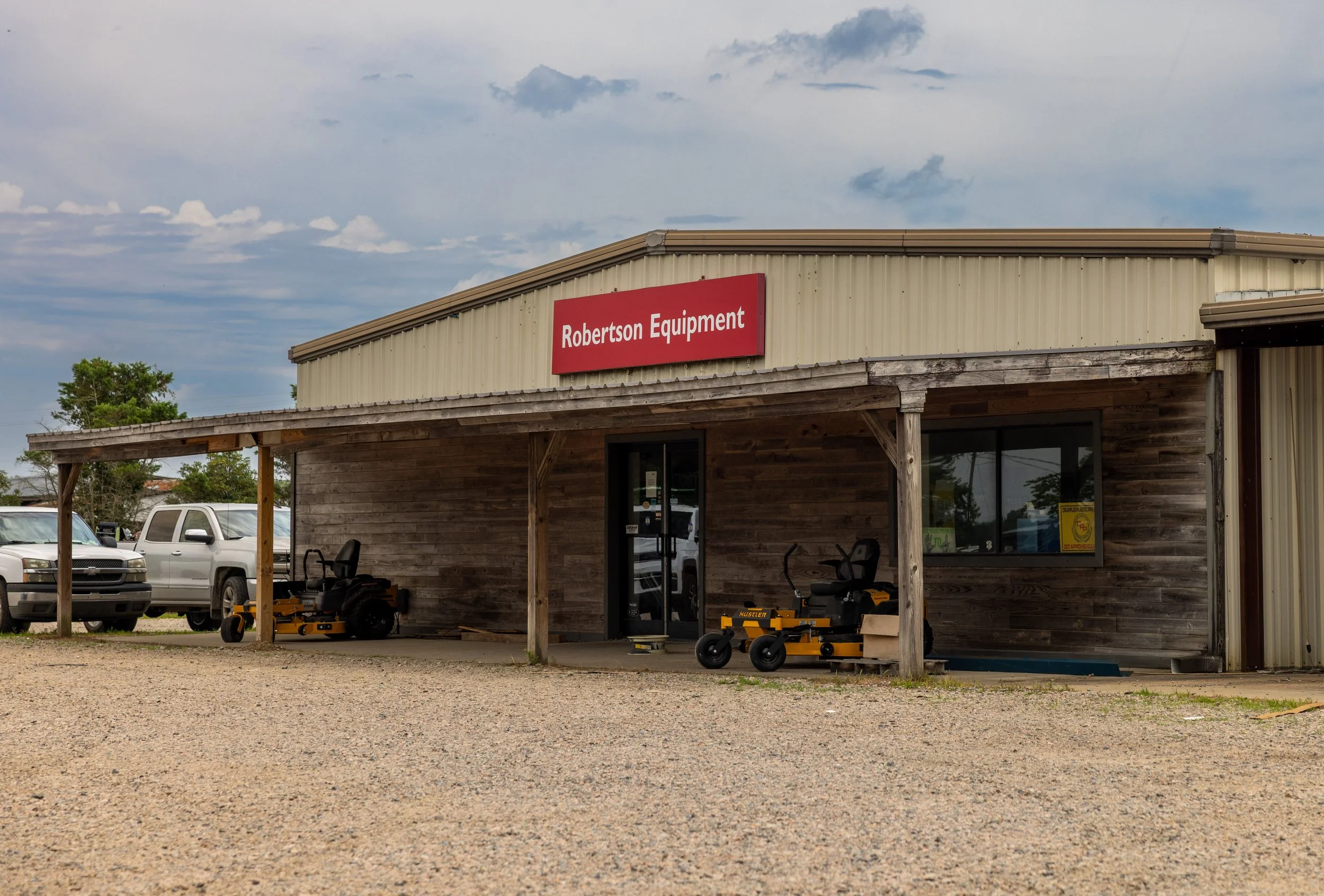 Front view of Robertson Equipment store with metal siding, wooden accents, and outdoor equipment and lawn mowers displayed outside. Several vehicles are parked nearby, under a partly cloudy sky.
