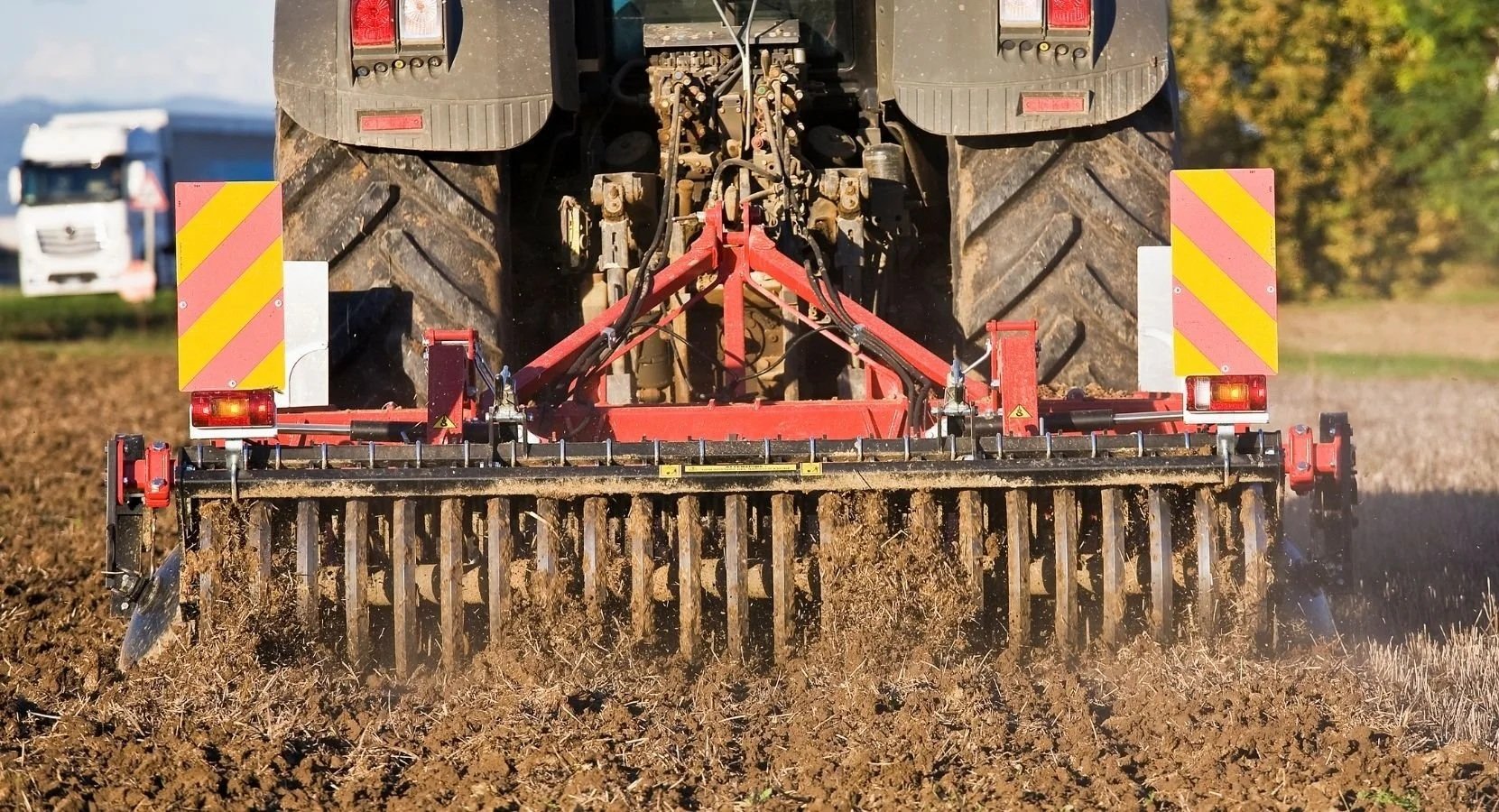 Close-up view of a tractor in a field, equipped with a red tillage implement, working on the soil during daytime with trees in the background.