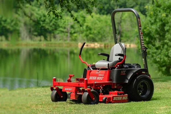 Red Bush Hog zero-turn lawn mower on a grassy area near a pond with trees in the background.