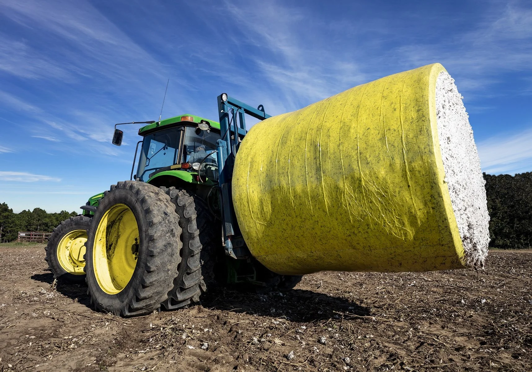 A green tractor with large yellow wheels and a yellow roller attachment on the front is on a dirt field under a blue sky with wispy clouds.