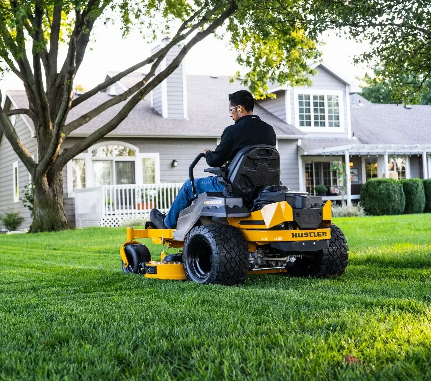 A man is sitting on a yellow Husqvarna Husqler riding lawn mower, mowing a well-maintained green lawn in a residential backyard with a two-story house, trees, and bushes.