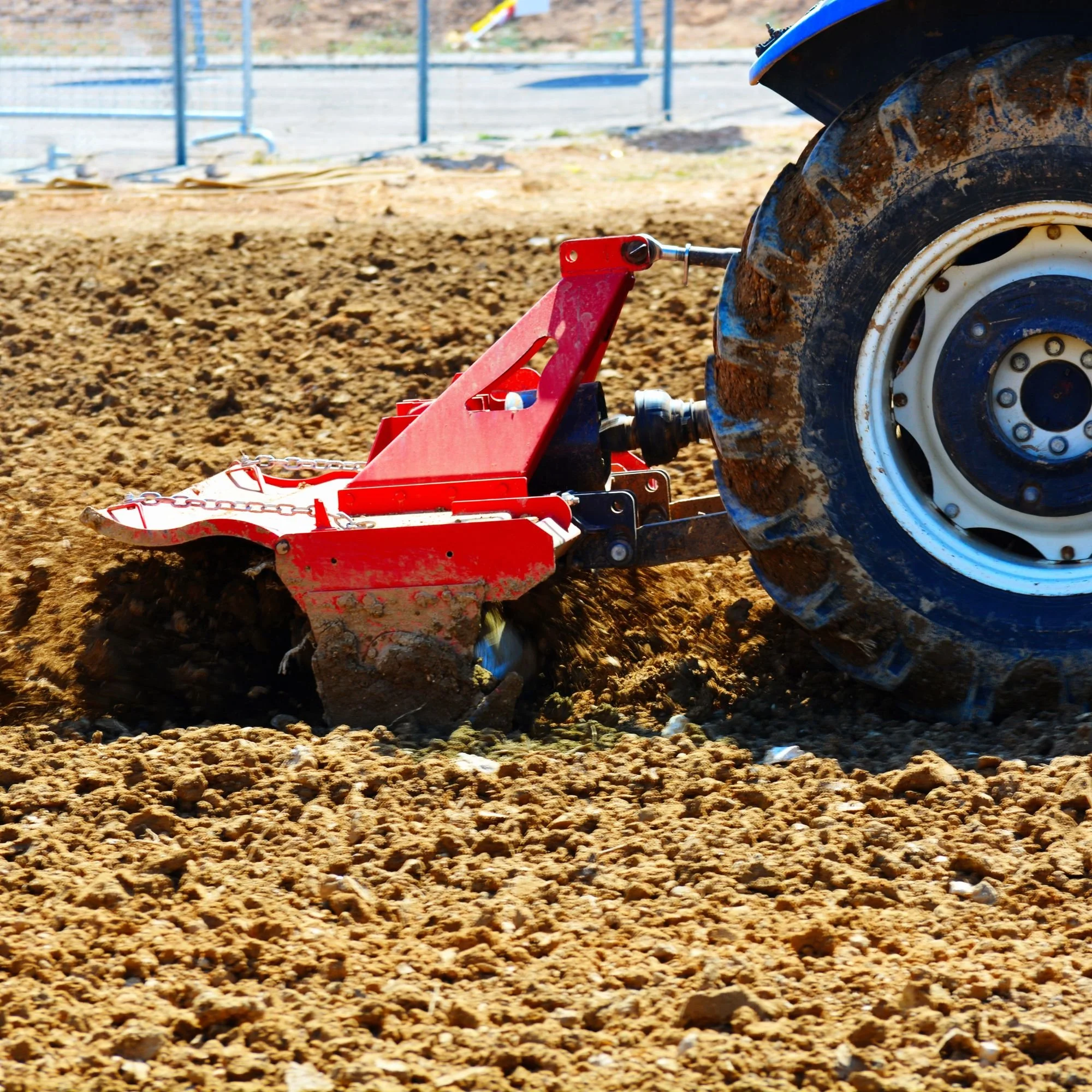 A tractor with muddy tires plowing a field with soil and a metal fence in the background.