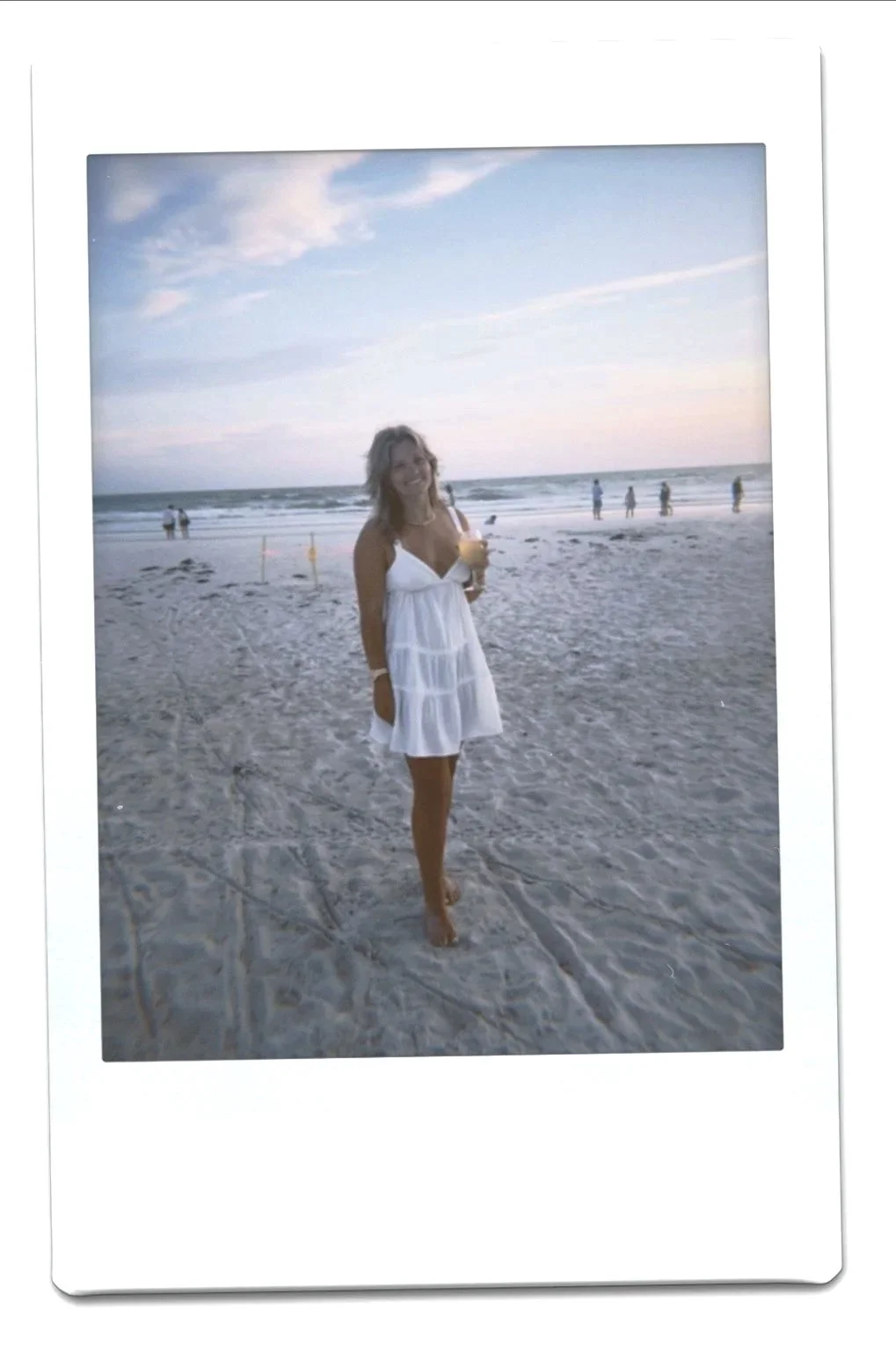 A woman in a white dress smiling while holding a drink and walking barefoot on a sandy beach, with the ocean and a few people in the background during sunset.
