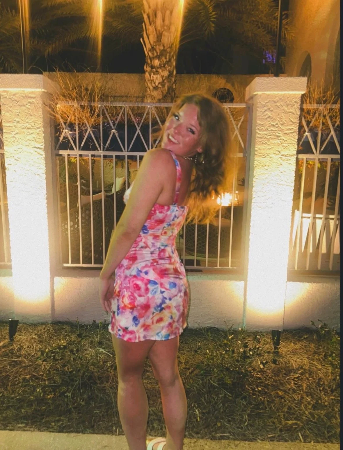 A woman in a colorful floral dress smiling and posing outdoors at night with a decorative metal fence and palm trees illuminated by warm lights in the background.