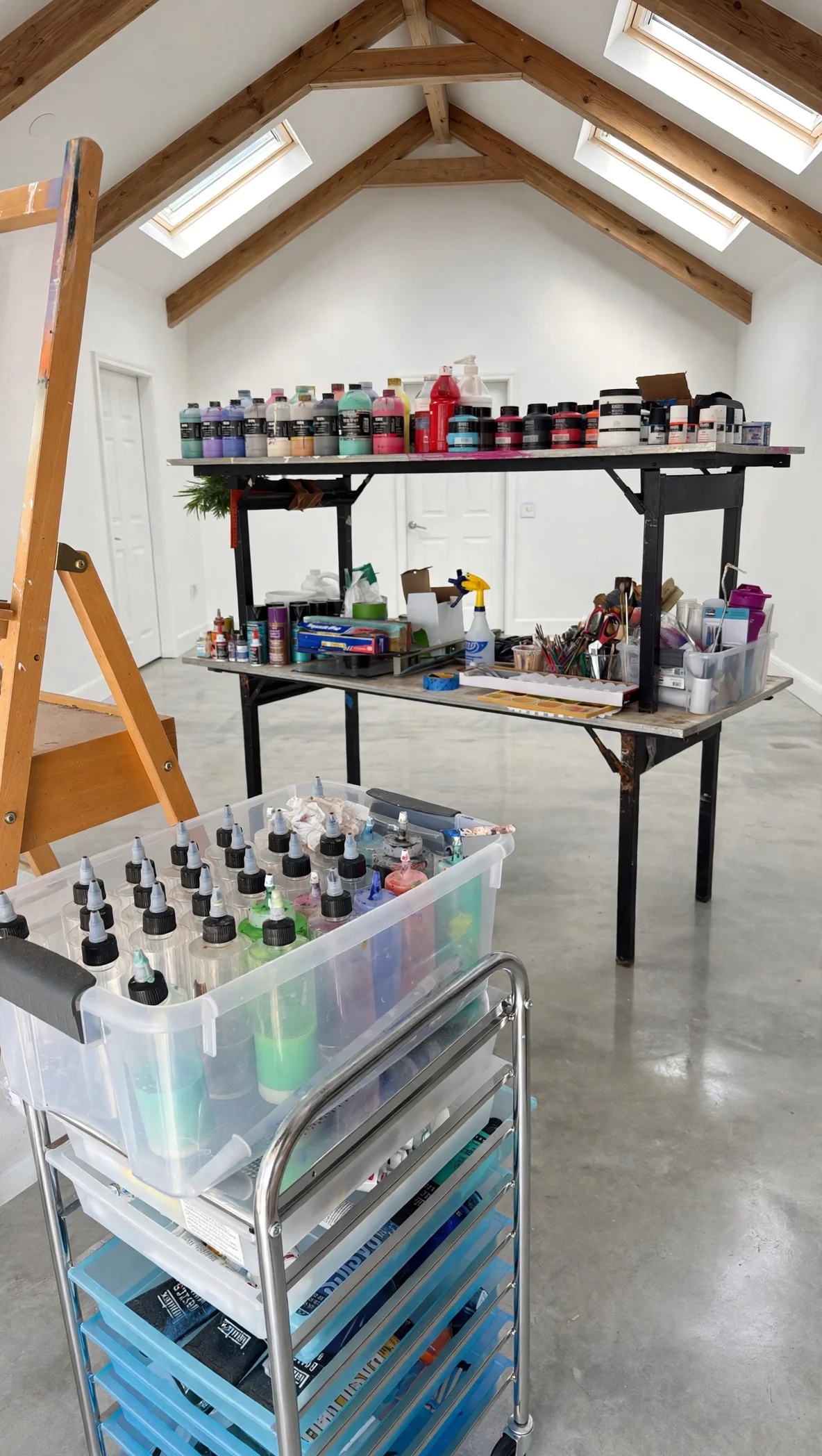 A room with a high, sloped ceiling featuring wooden beams and three skylight windows. There is a work table with various art supplies, including bottles of paint, brushes, and tools. A plastic cart with multiple trays of inks or dyes is in the foreground, and a ladder is partially visible on the left side.