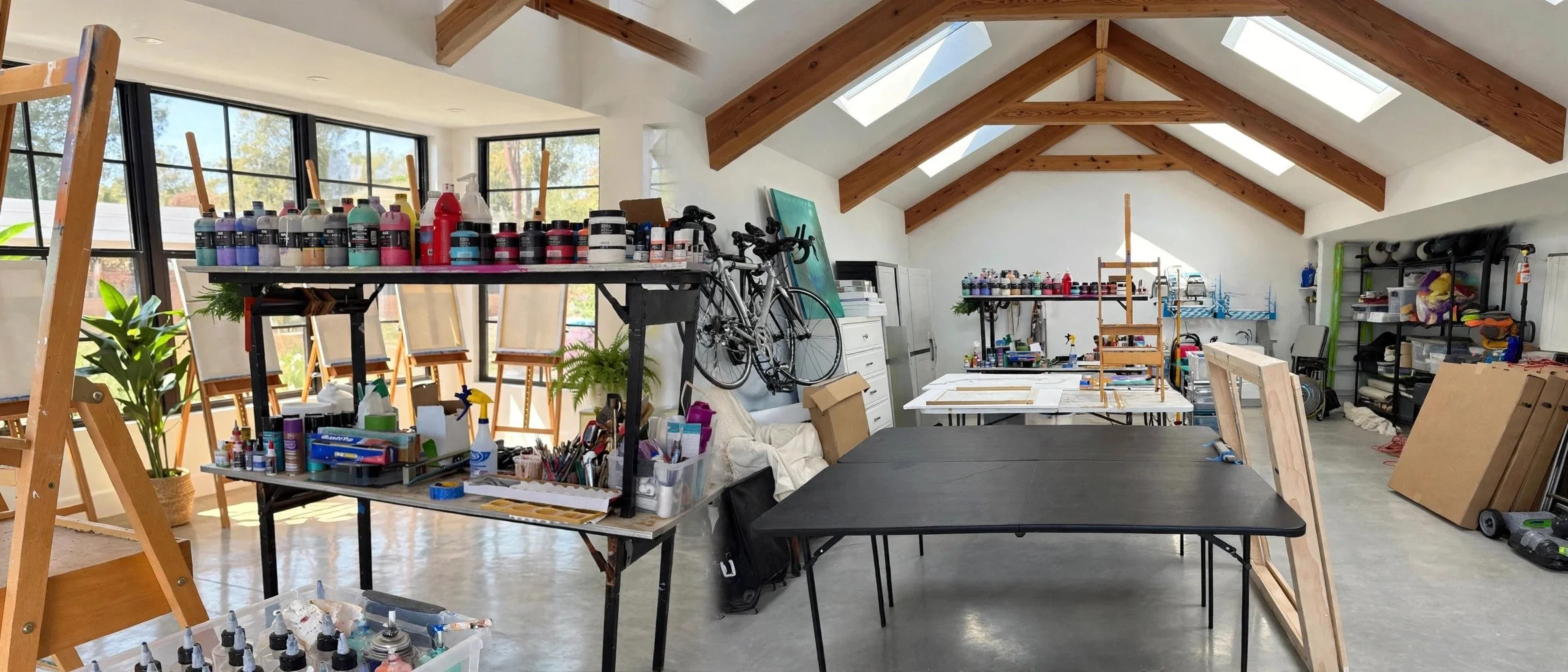 An art studio with work tables, shelves filled with art supplies, bicycles on the wall, and a sloped ceiling with skylights, illuminated by natural light.