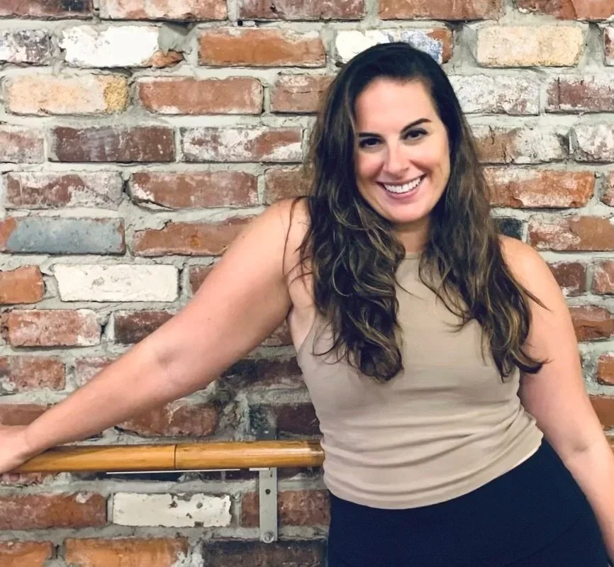 A smiling woman with long, wavy dark hair and light skin, wearing a sleeveless beige top and black pants, leaning against a wooden ballet barre in front of a brick wall.