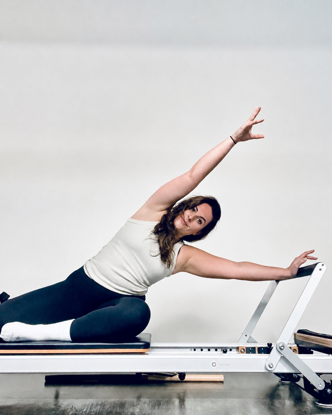 Woman doing a side stretch exercise on a reformer Pilates machine, smiling, with a plain white background.
