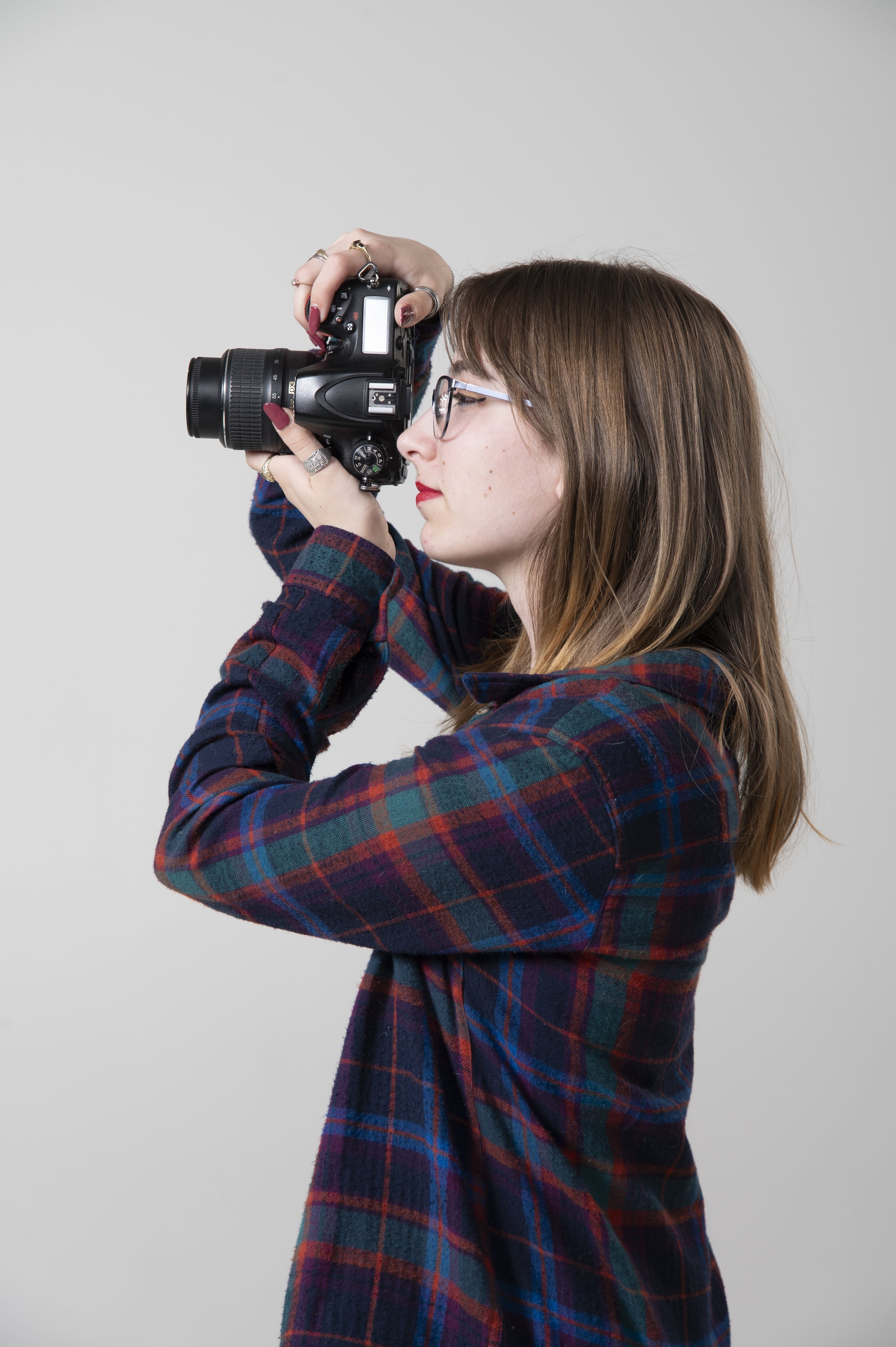 A woman with long brown hair, wearing glasses and a plaid shirt, holds a camera up to her face as if taking a photo, against a plain gray background.