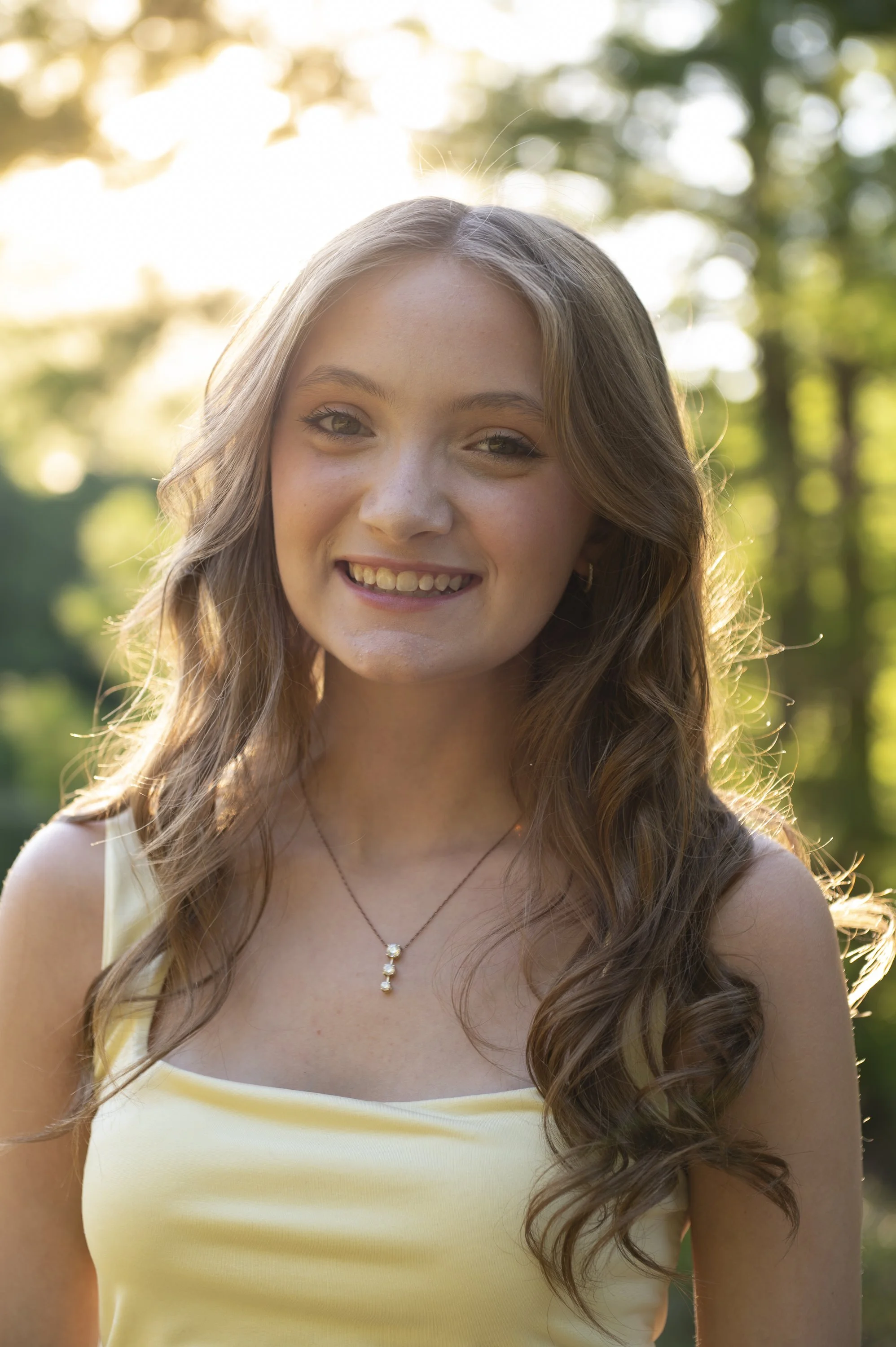 Close-up of a young woman with long, curly brown hair, smiling in a park with sunlight filtering through trees in the background.
