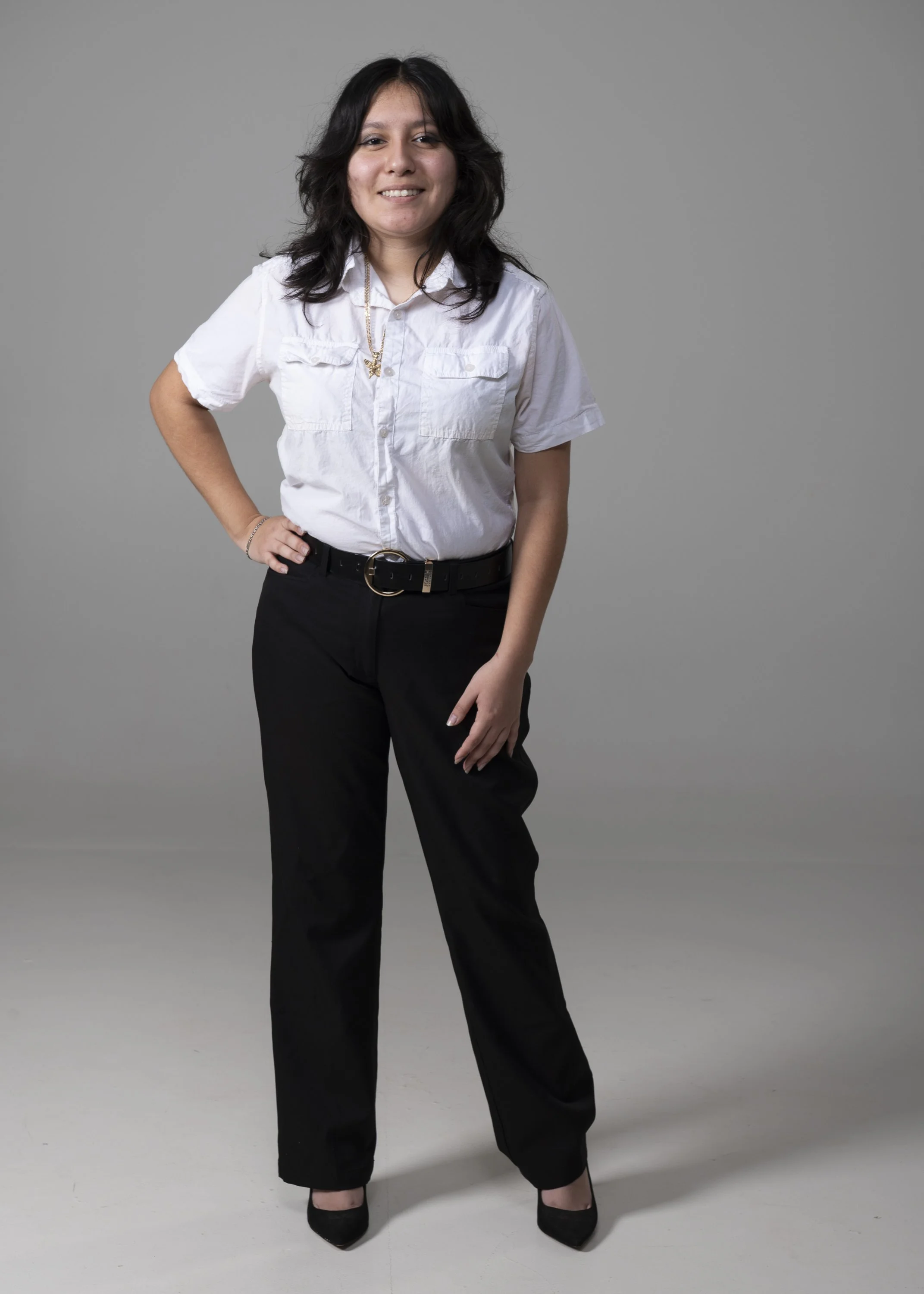 A woman with dark curly hair, wearing a white button-up shirt, black trousers, black heels, a gold necklace, and a bracelet, standing with her hand on her hip against a plain gray background.