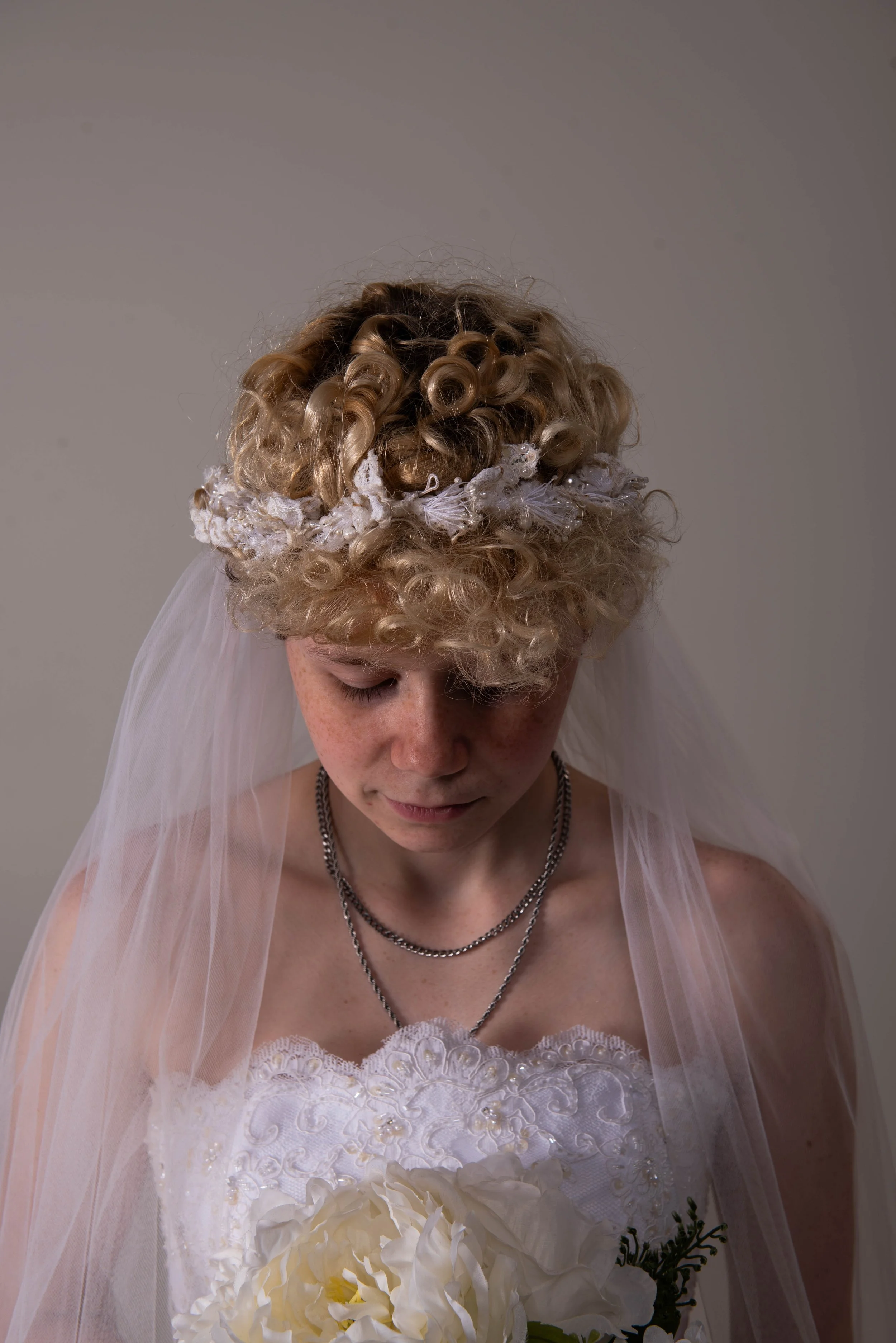 A woman with light skin and curly blonde hair, wearing a white wedding dress with lace details and a veil, holding a bouquet of white flowers and greenery, with her eyes closed.