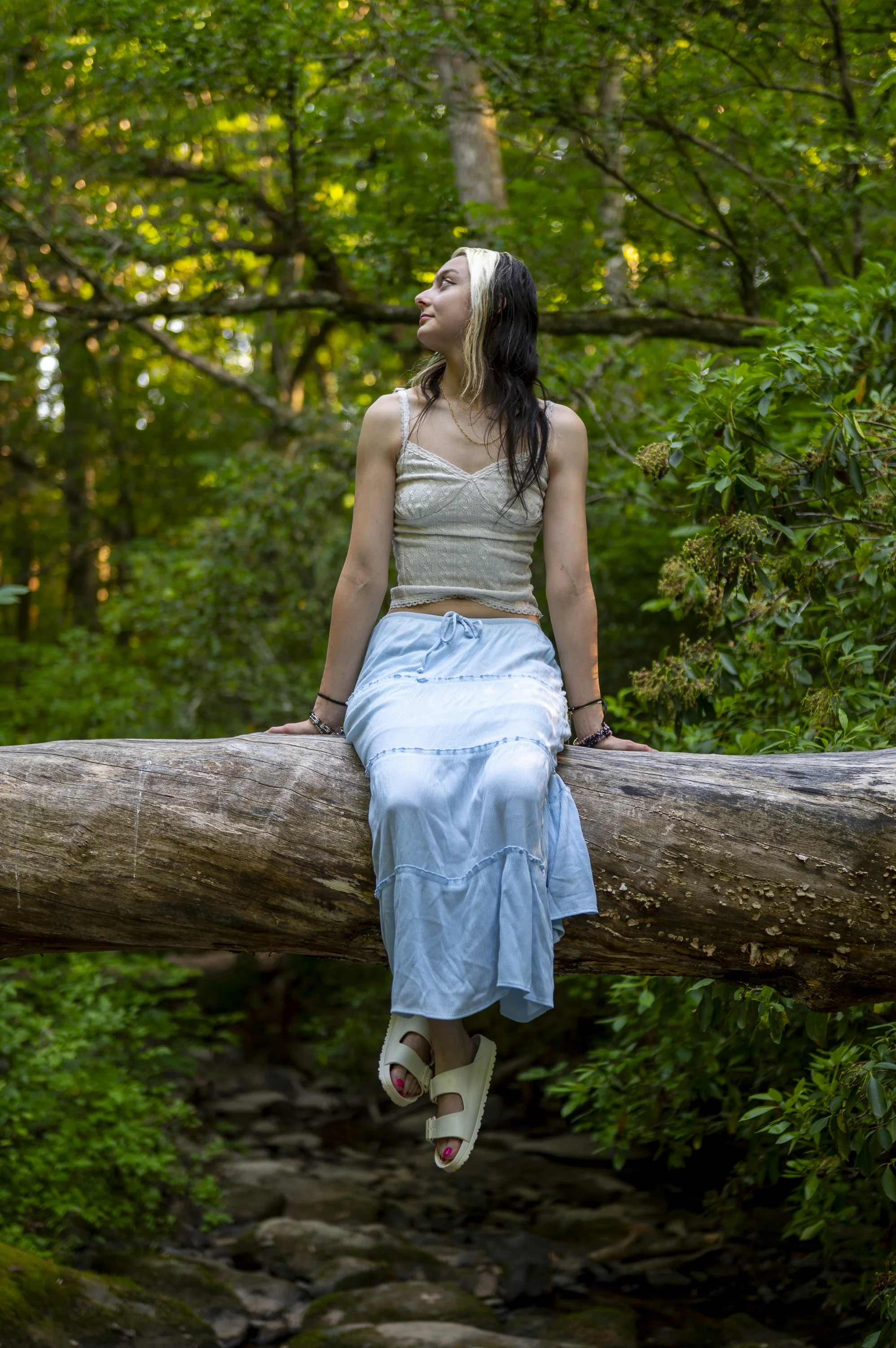 A young woman with long dark hair with a blonde streak, wearing a beige lace crop top, a white skirt, and white platform sandals, sitting on a large fallen tree trunk in a lush green forest, looking to her left.