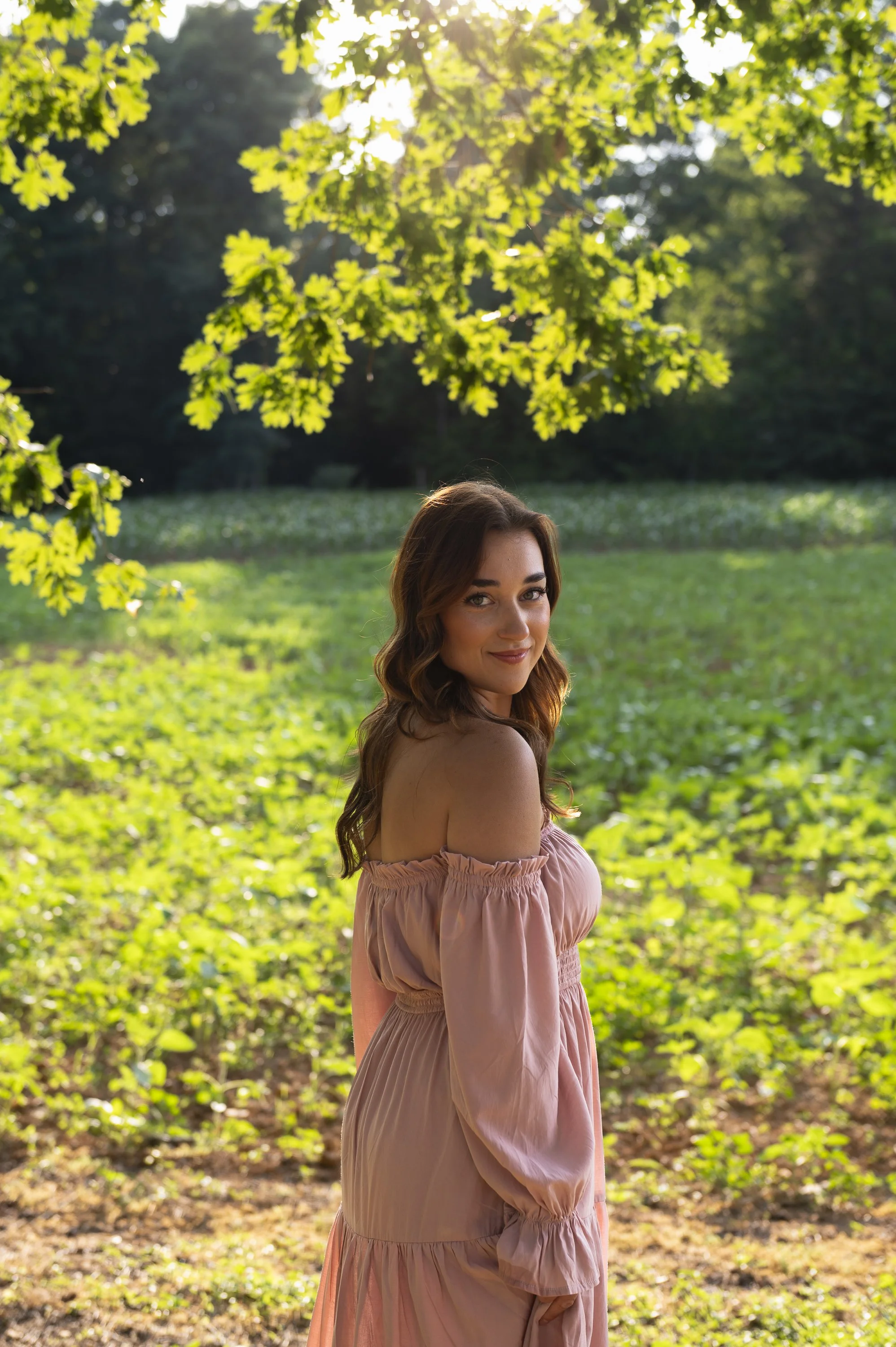 Young woman with wavy brown hair smiling outdoors in a pink off-shoulder dress, standing in a green park with trees and grass, sunlight filtering through the leaves.