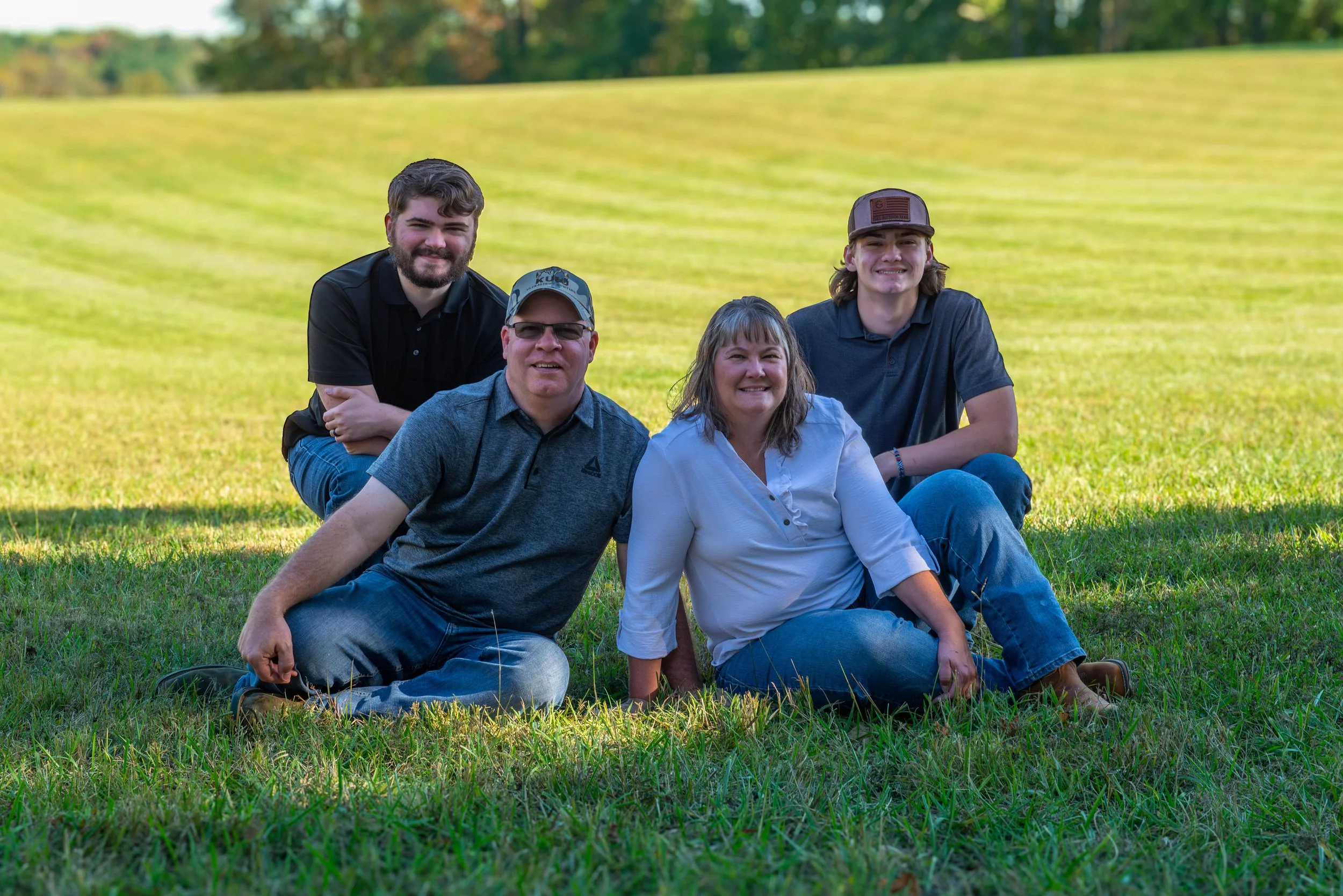 A family of five sitting outdoors on a grassy field, smiling at the camera. The background shows a green, slightly sloped landscape with trees.