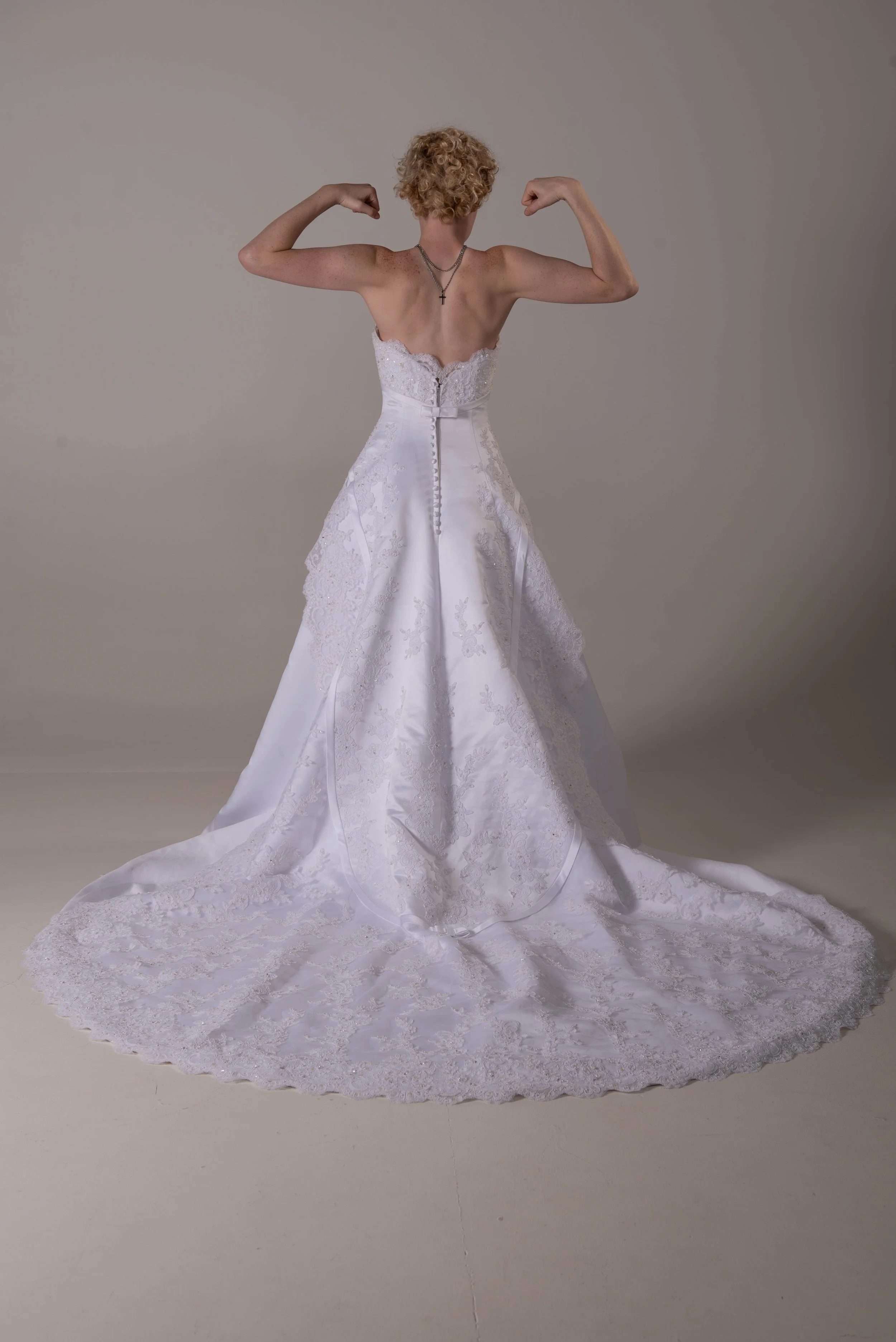 A woman with curly blonde hair wearing a white wedding dress, flexing her arms to show muscle, standing against a plain gray background.
