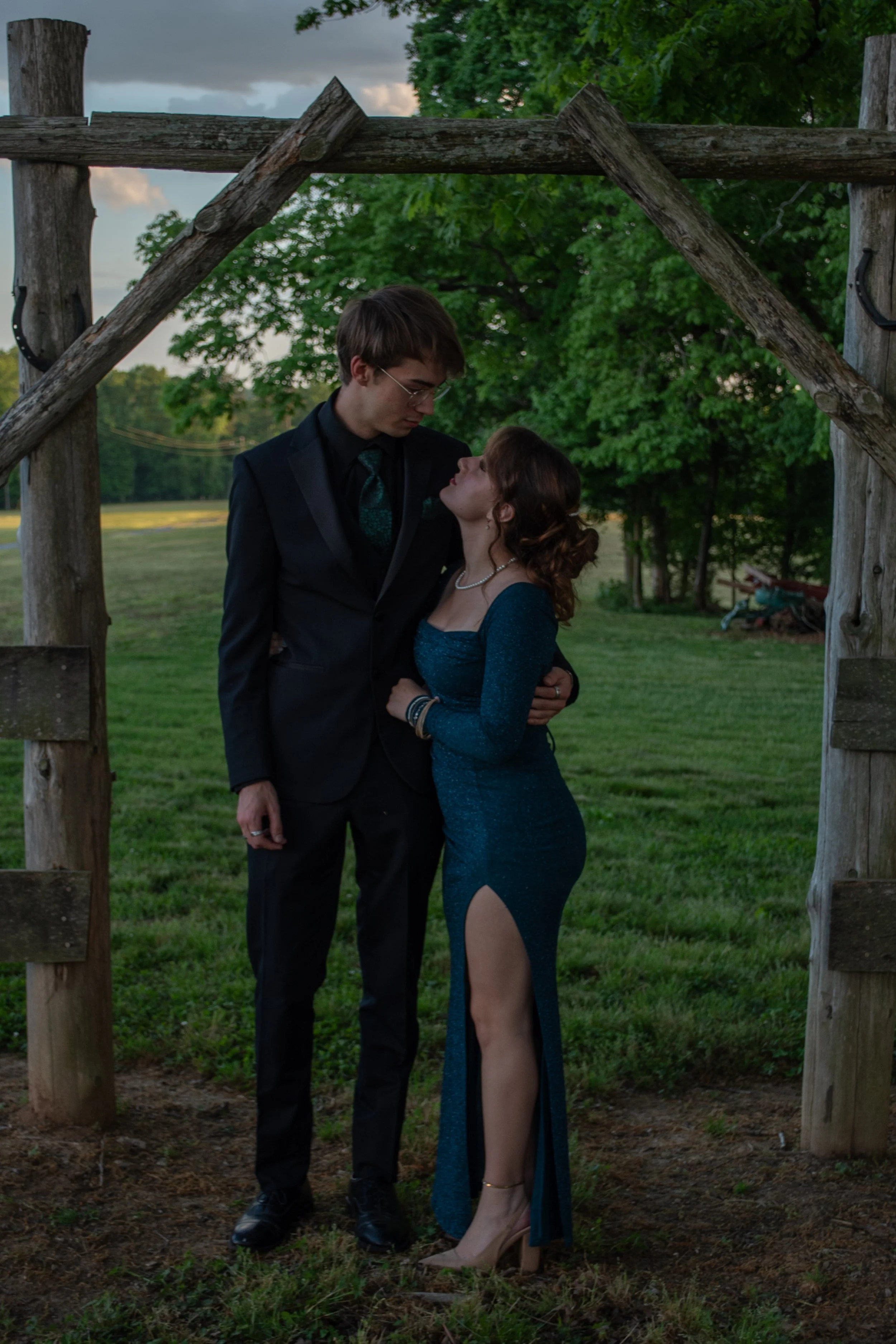 A young man in a black suit and a young woman in a glittery green dress stand close together under a rustic wooden archway outdoors, with green trees and a cloudy sky in the background.