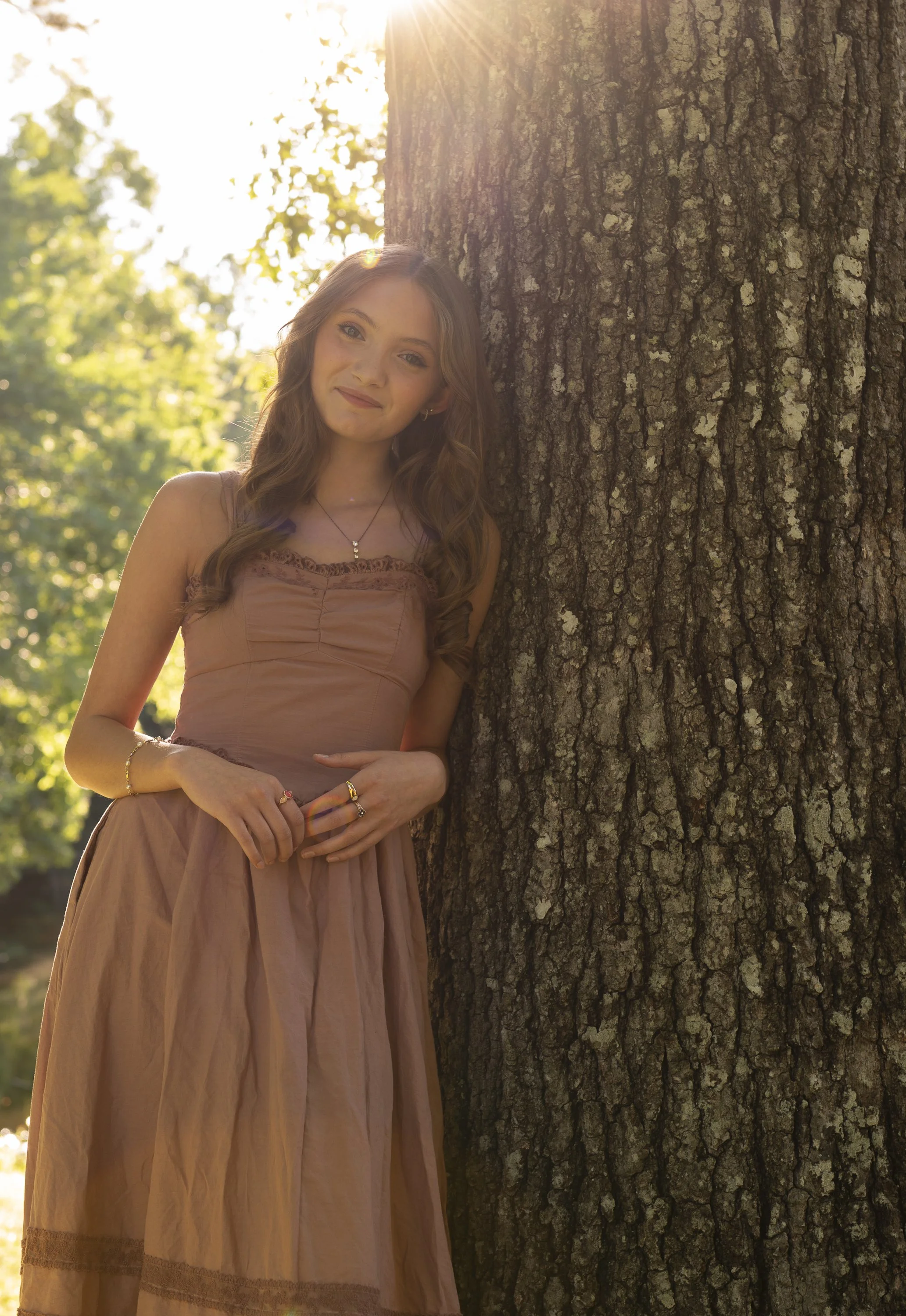 A young woman with long wavy hair wearing a sleeveless dress with lace details, standing outdoors next to a large tree with textured bark, illuminated by sunlight in a park with green foliage in the background.