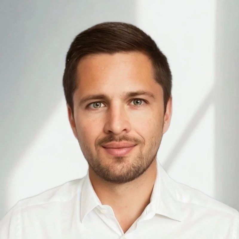 A portrait of a young man with short brown hair and a beard, wearing a white collared shirt, standing in front of a light background.