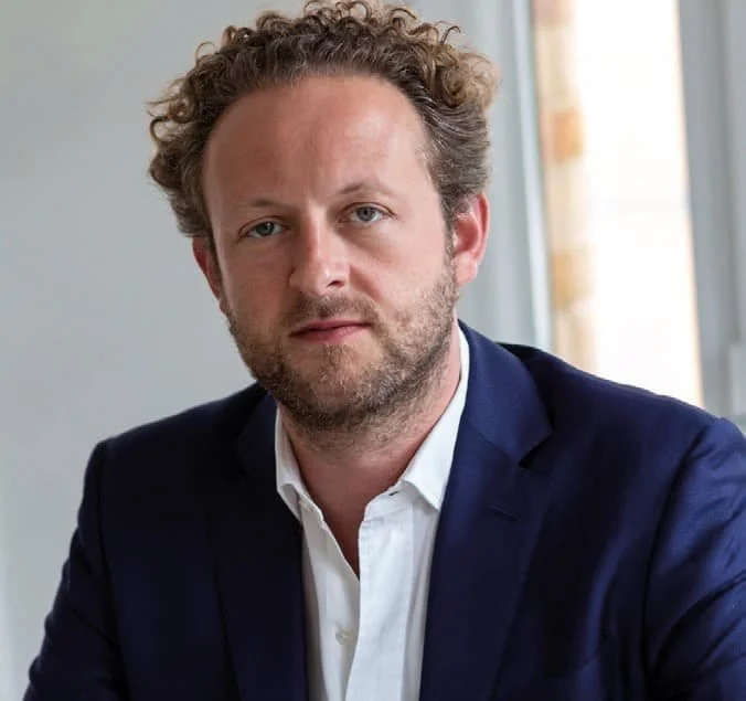 A man with curly hair and a beard wearing a navy blazer and white shirt, sitting in front of a window with natural light.