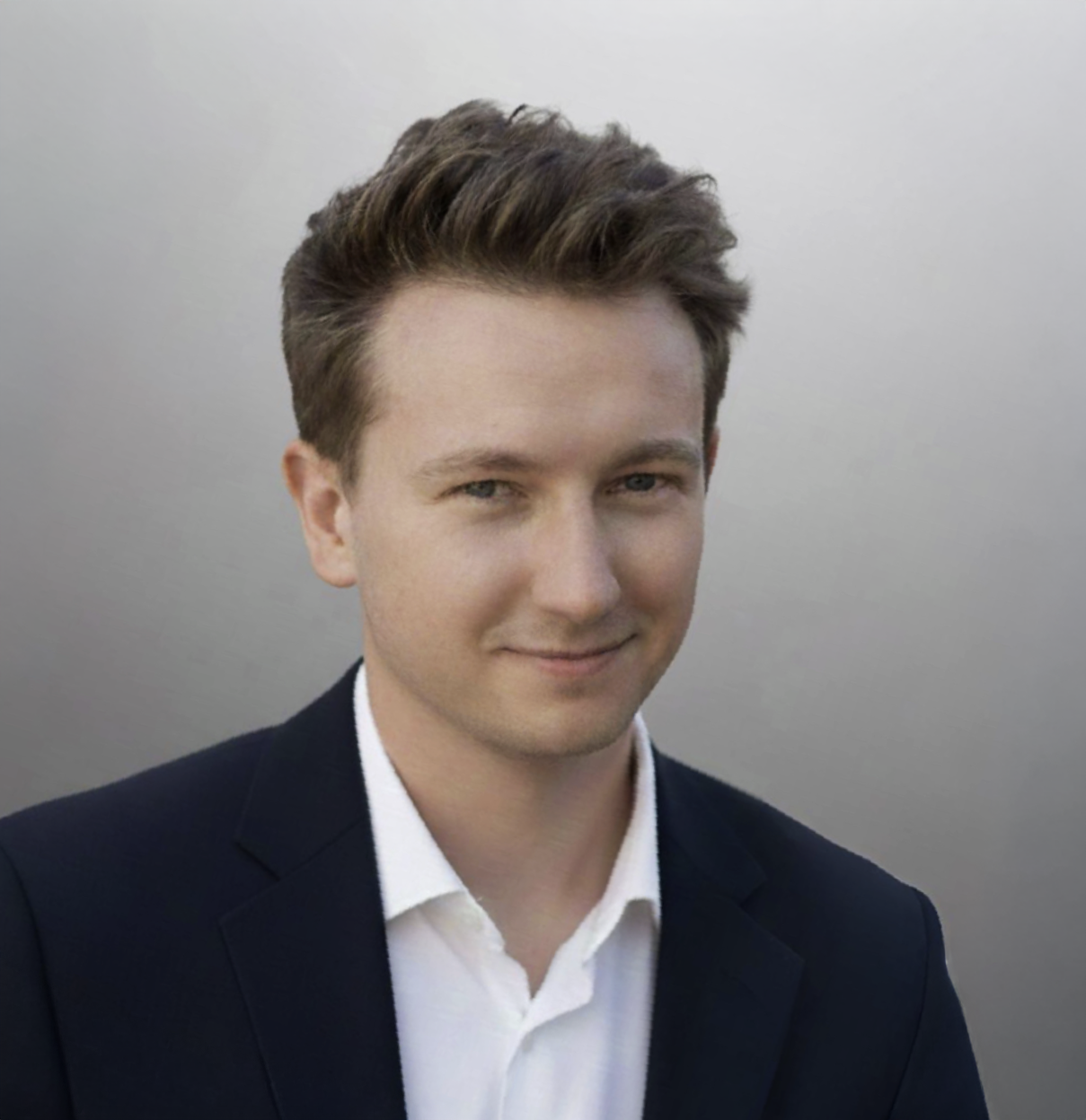 A young man with brown hair and blue eyes dressed in a black suit and white shirt, smiling slightly against a plain gray background.