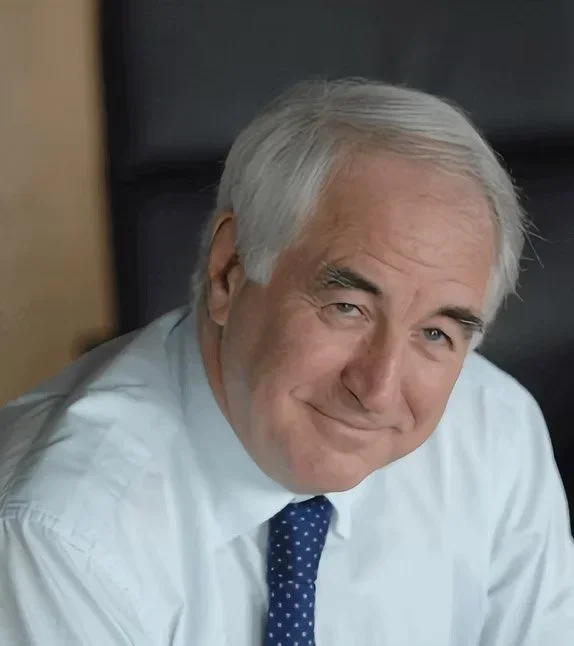 A senior man with gray hair smiling and wearing a white shirt with a blue polka dot tie, sitting at a desk in an office.
