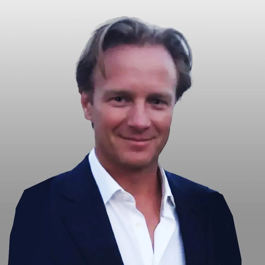 A portrait of a man with dark hair, wearing a white shirt and navy blazer, smiling against a plain light gray background.