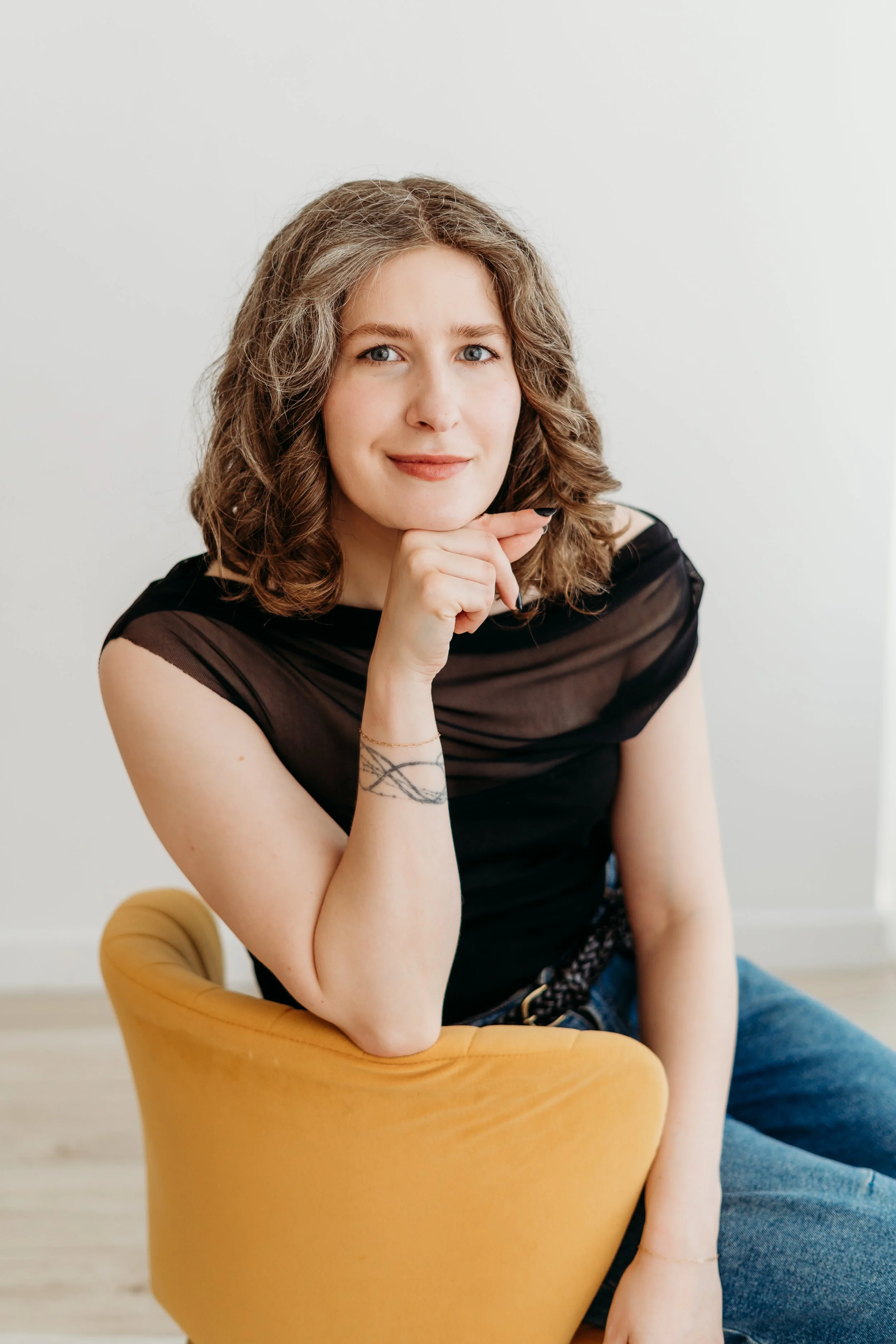 a woman who works as a beauty product developer is wearing a black shirt and jeans and sitting on a yellow chair. she is smiling, with her chin rested on her hand