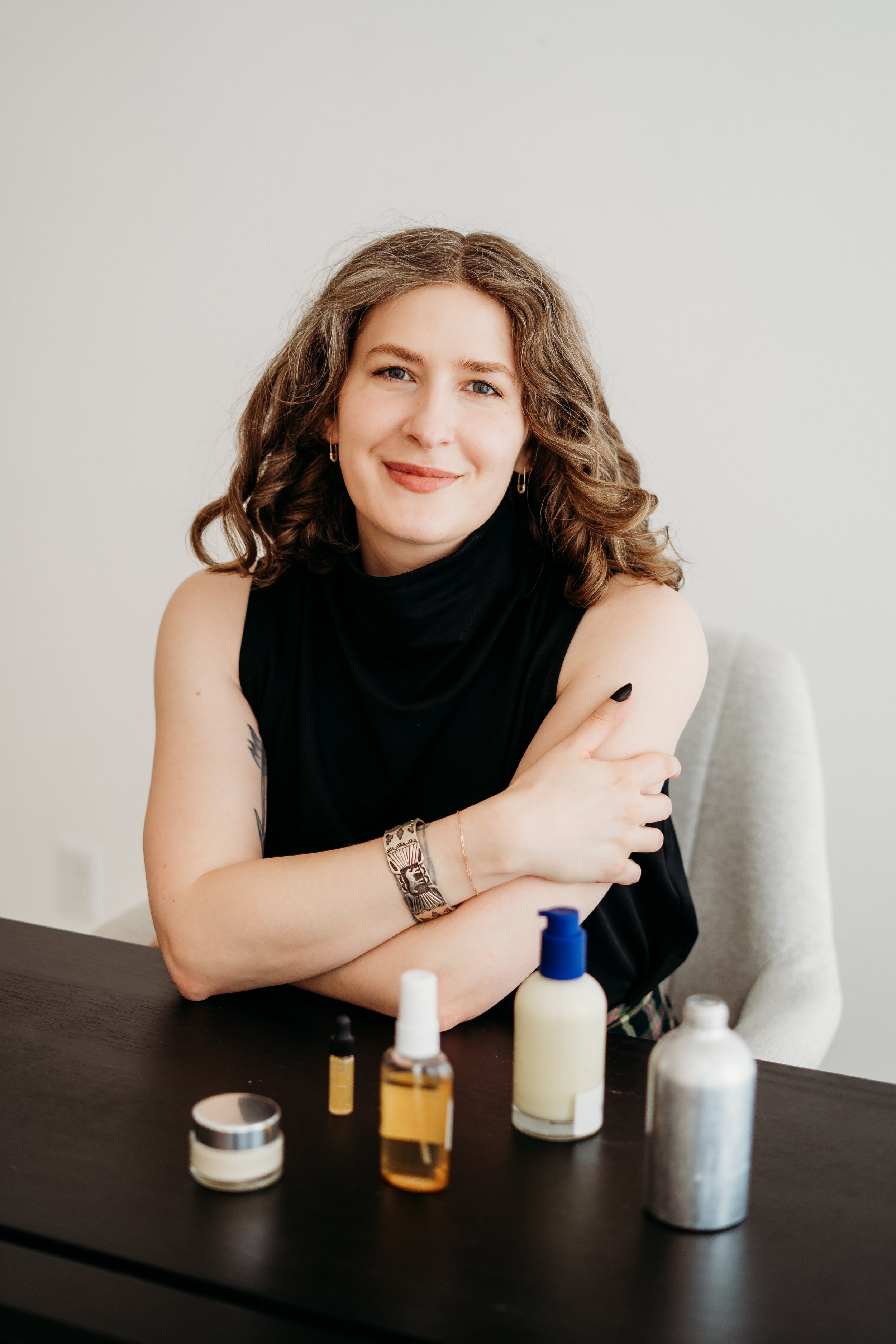 A woman in a black sleevess shirt sits at a desk, smiling. On the desk are beauty products and packages she developed.