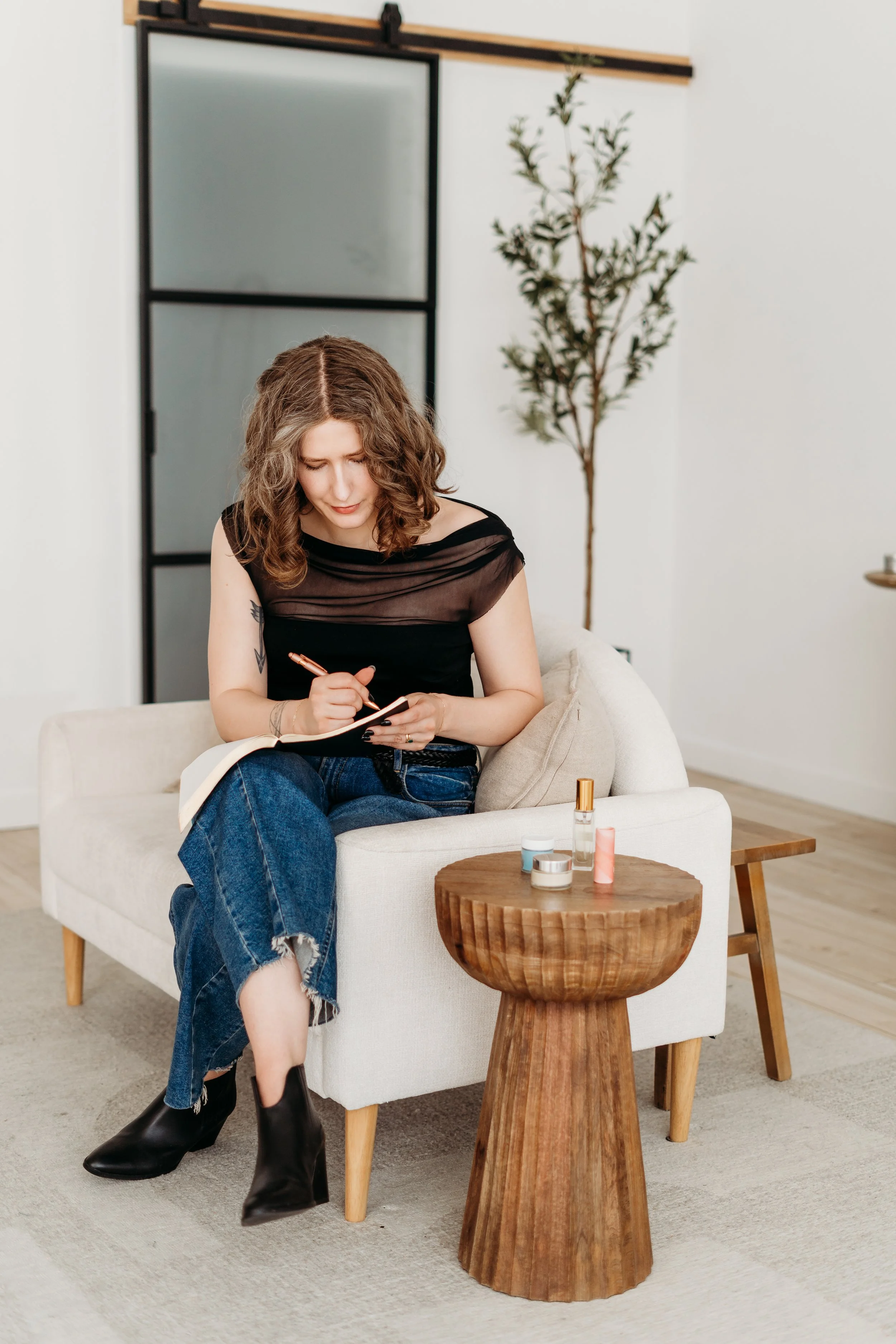 A woman with curly brown hair, wearing a black top and ripped jeans, sits on a white armchair, writing in a notebook. There are several small bottles and containers of skincare or beauty products on a wooden side table next to her. The background features a frosted glass sliding door and a tall indoor plant.