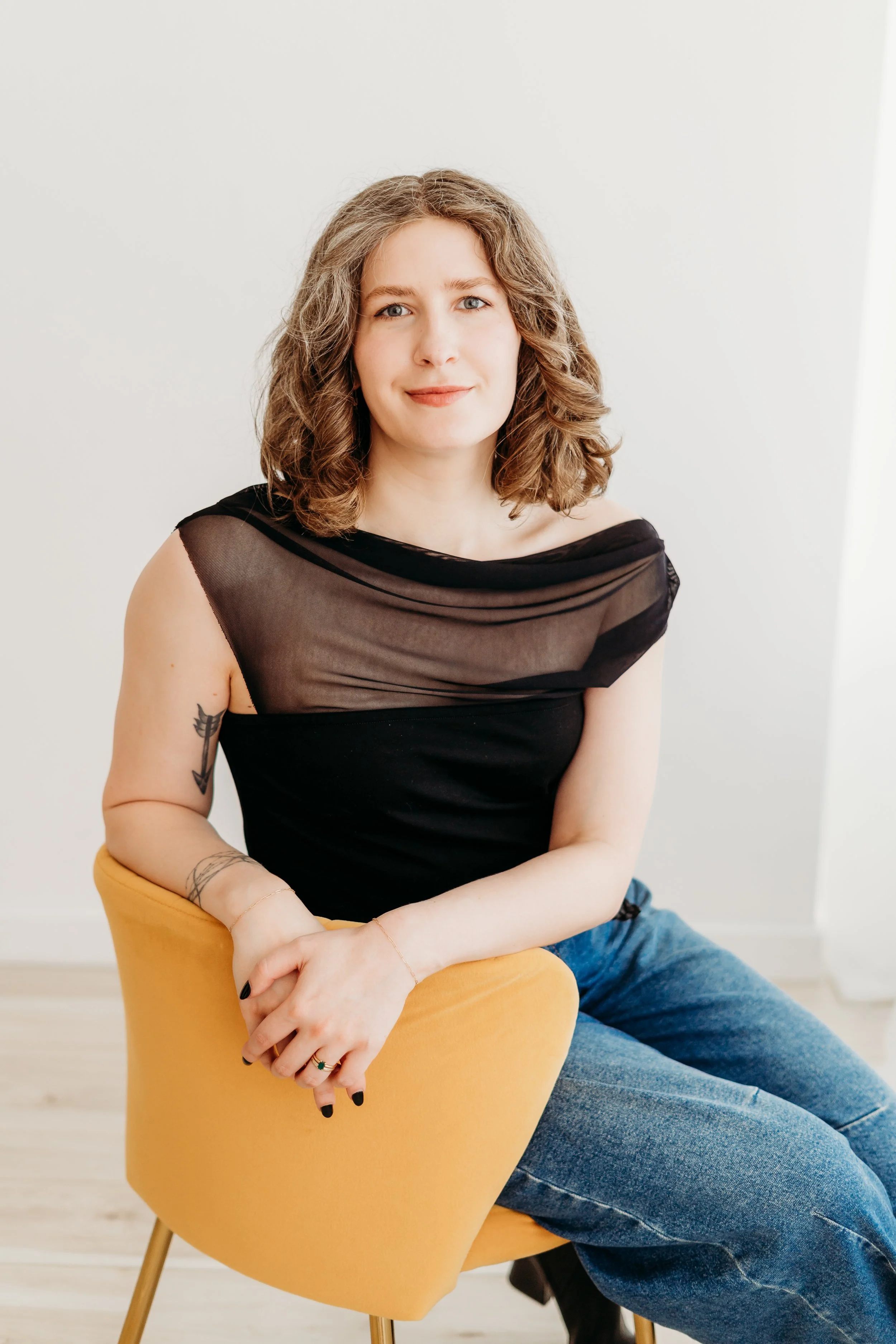 A woman with curly brown hair and blue eyes sitting on a mustard yellow chair, wearing a black sheer top over a black tank top and blue jeans, smiling softly, with a white wall background.
