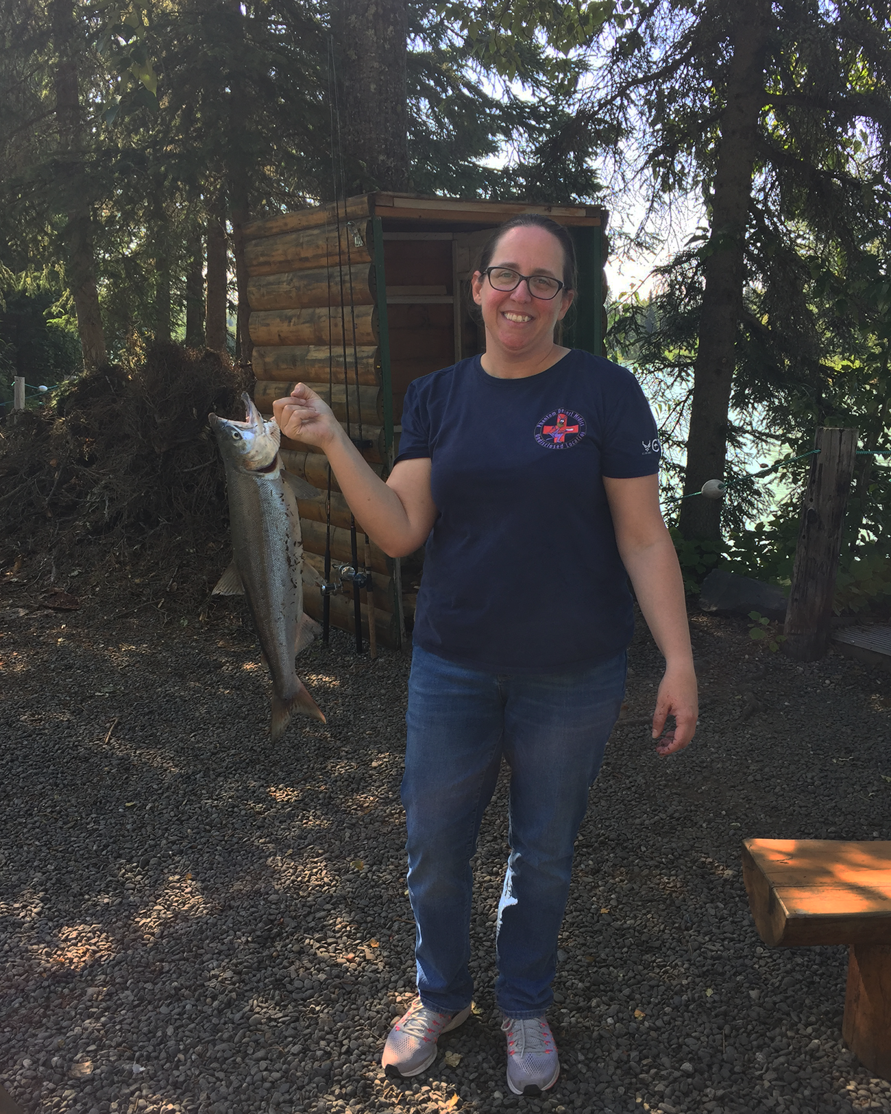 Doctor Liz is standing in front of a cabin by a wooded body of water. She is smiling into the camera and looks at ease. In her hand, she holds a large salmon she has caught fishing.