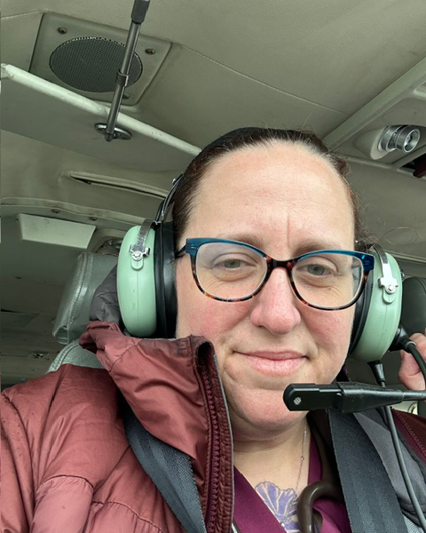 A more recent picture of Liz smiling softly into the camera, once again wearing a pilot's headset as she flies. She wears a warm looking rust colored puffy coat.