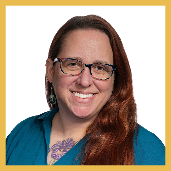 A portrait of Dr. Elizabeth Pietralczyk. She is smiling warmly into the camera. She has long red hair, multicolored glasses, and a tattoo of a purple iris can be seen above the neckline of her teal shirt.
