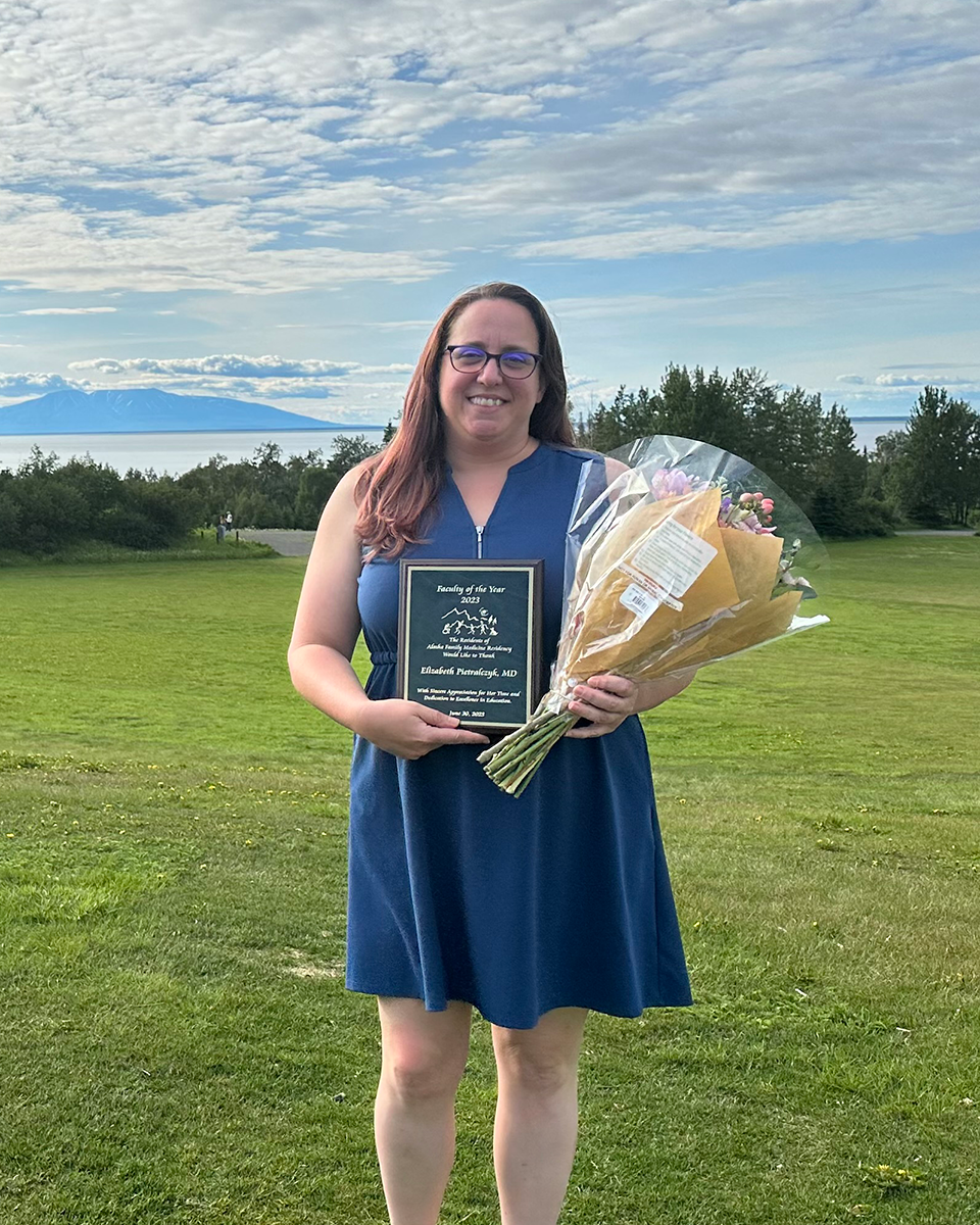 Doctor Liz smiles into the camera as she holds a Faculty of the Year award and a bouquet of flowers. She wears a blue dress, and stands in a sunny grass field.
