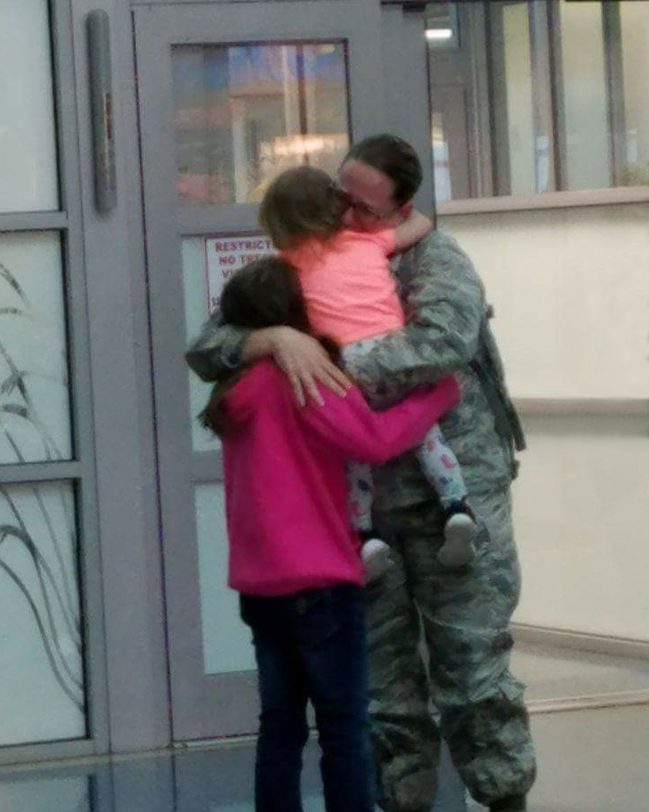 Doctor Liz is hugging her two children at a tearful, joyous reunion. She is in her fatigues, while her child and toddler are in colorful clothes.