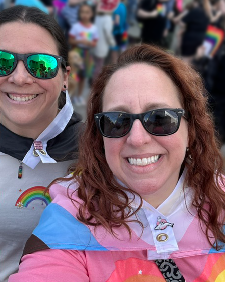 Doctor Liz and her partner smile into the camera, wearing sunglasses. They are decked out in Pride gear, Liz wearing the intersectional progress Pride flag around her shoulders. Behind them are blurred figures also attending a Pride event.