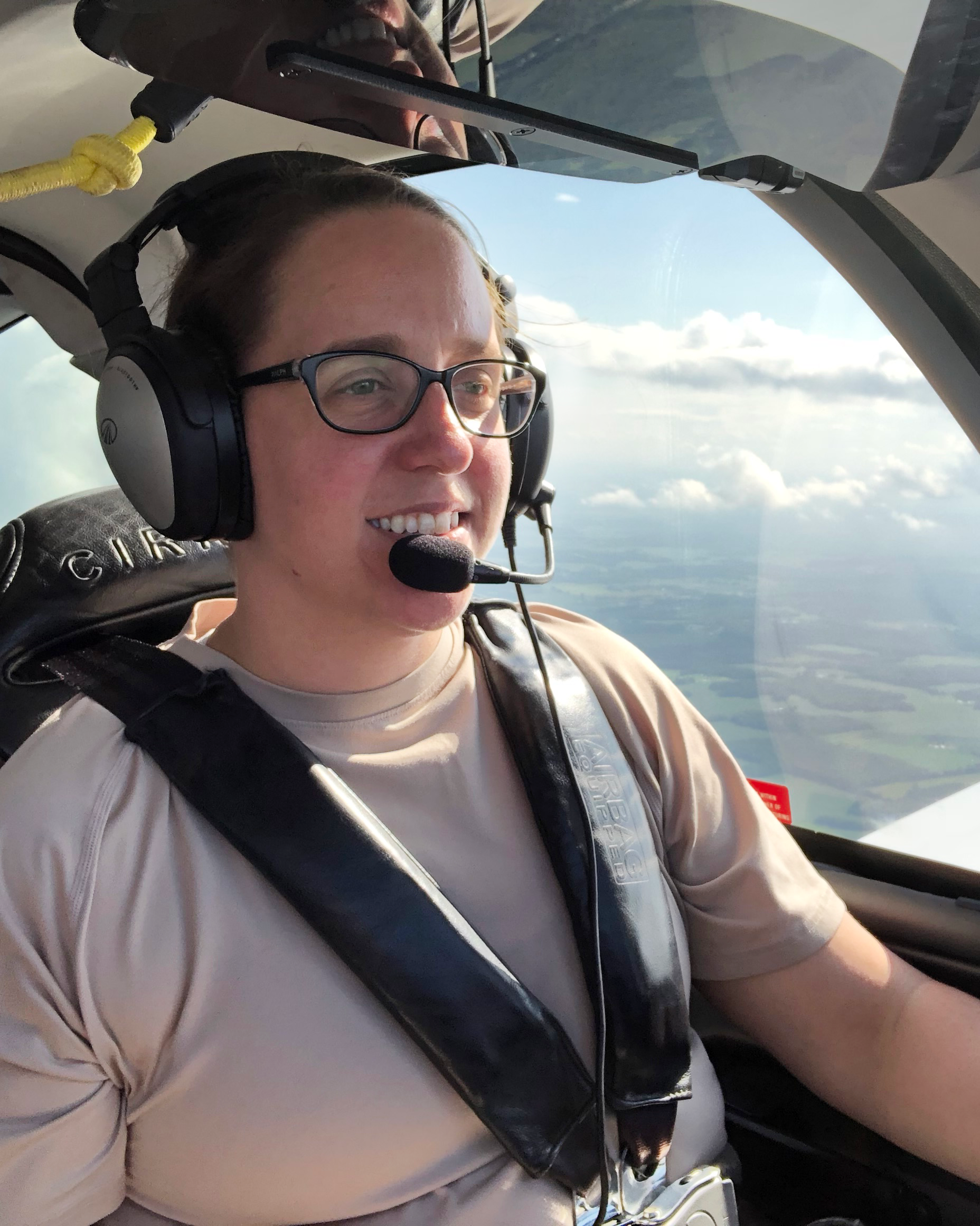 Doctor Liz sits in the cockpit, piloting a plane. She is wearing a headset and smiling.