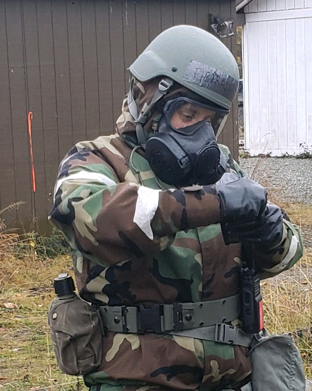 Doctor Liz is wearing her military fatigues, a helmet, and a respirator mask with goggles. She has a canteen on her hip and appears to be focused on something in her hands.
